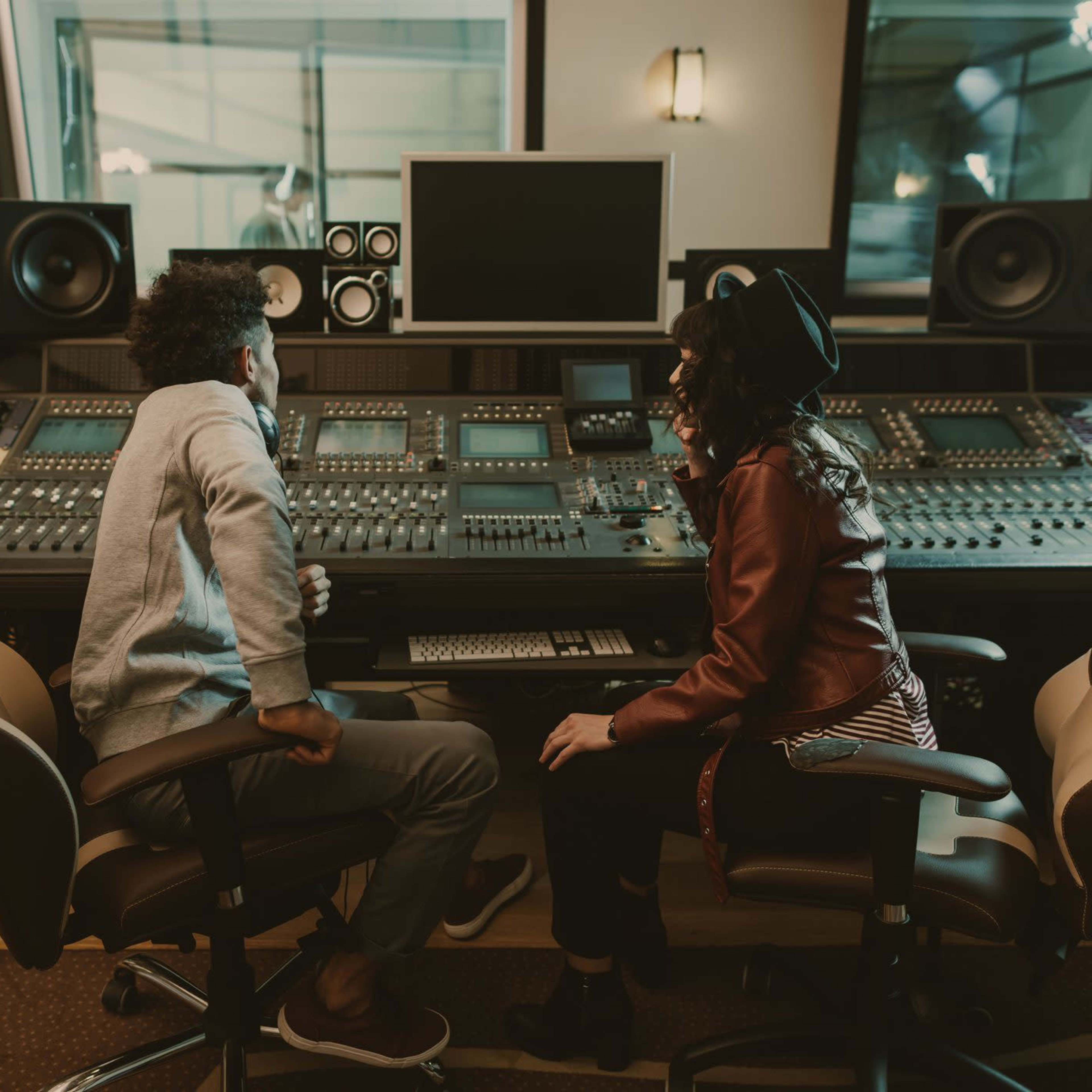 Two individuals are seated in front of a large music mixing console in a recording studio.