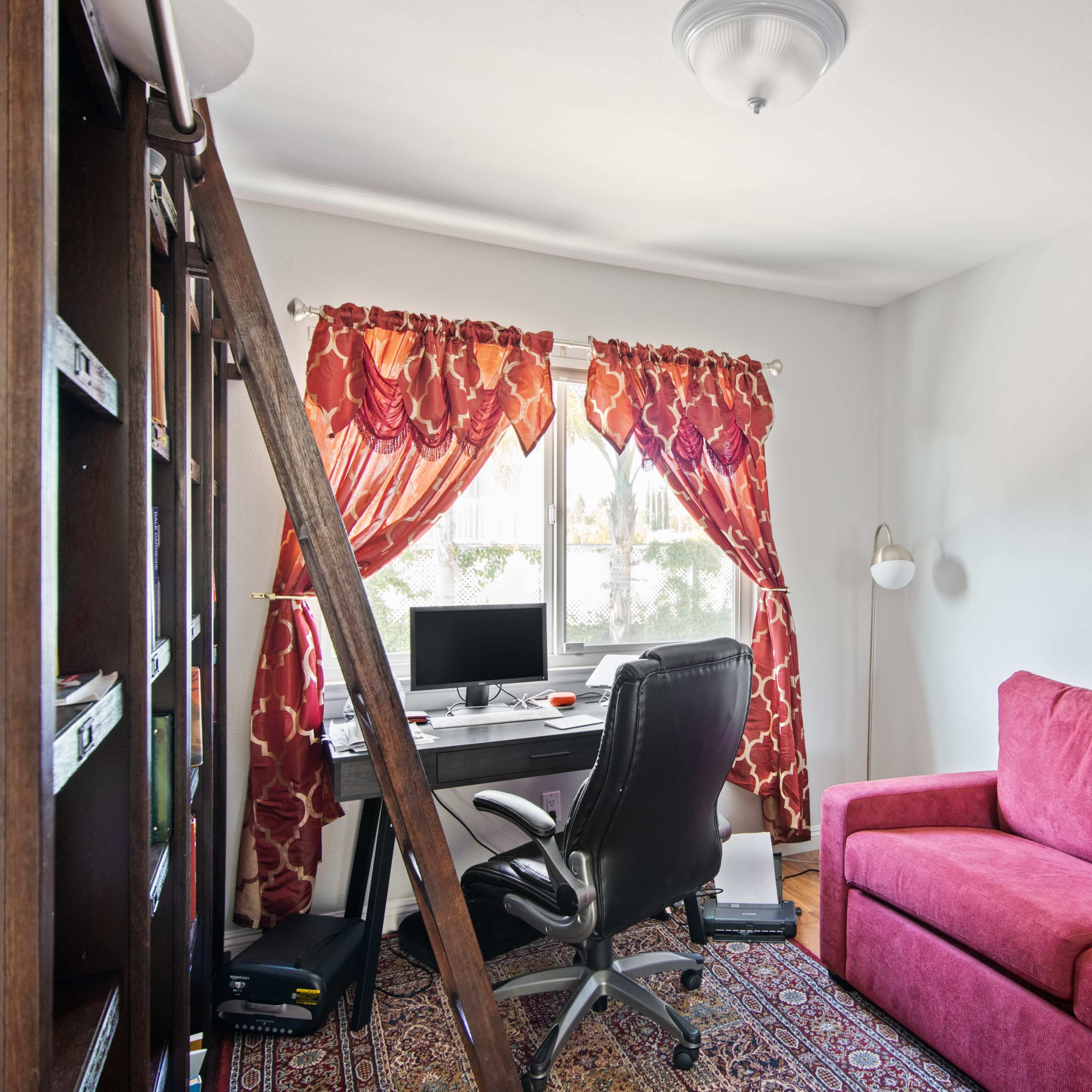 A small room features a desk with a computer, a black office chair, a red sofa, and large windows adorned with vibrant curtains.