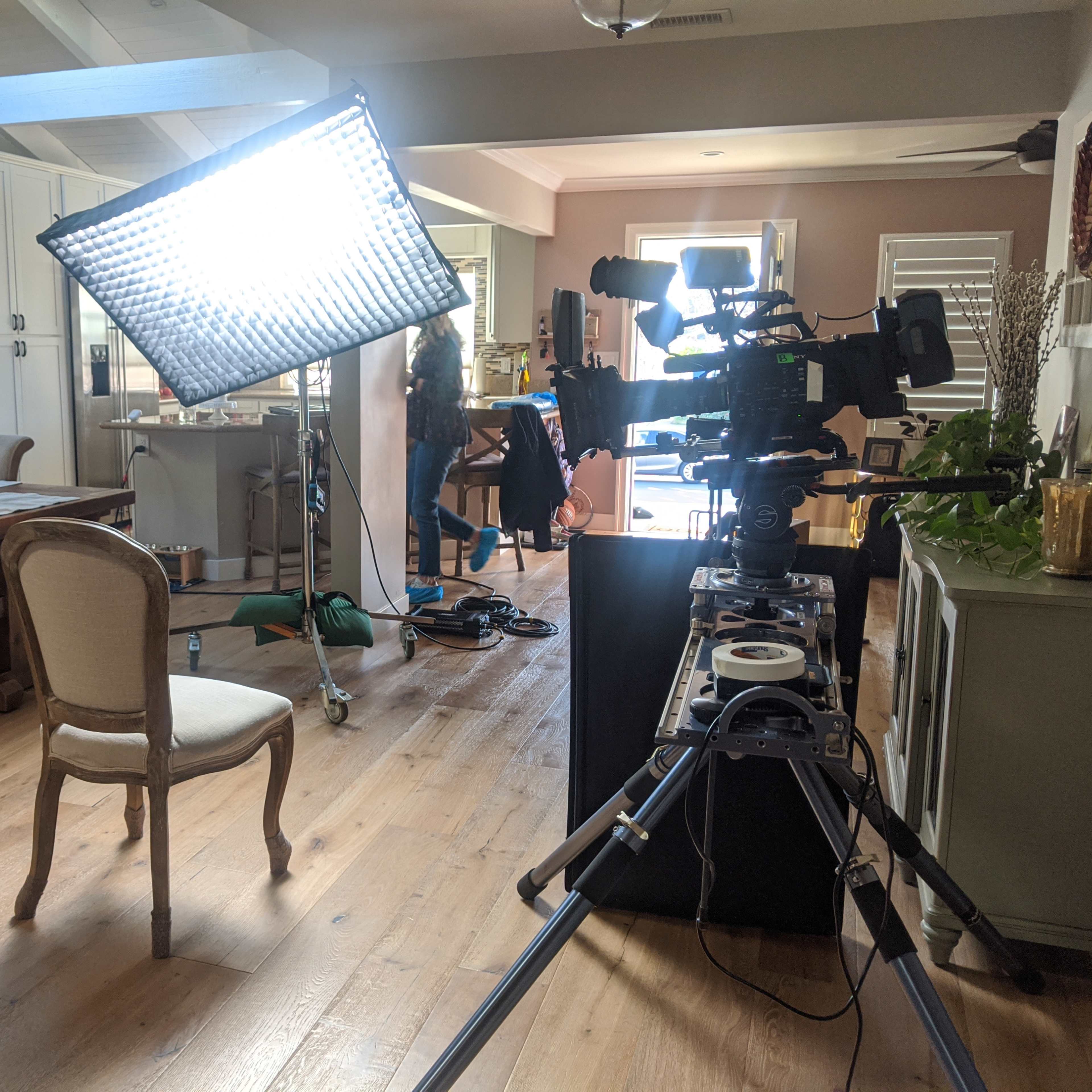 A video camera and lighting setup are positioned in a well-furnished kitchen and dining area.