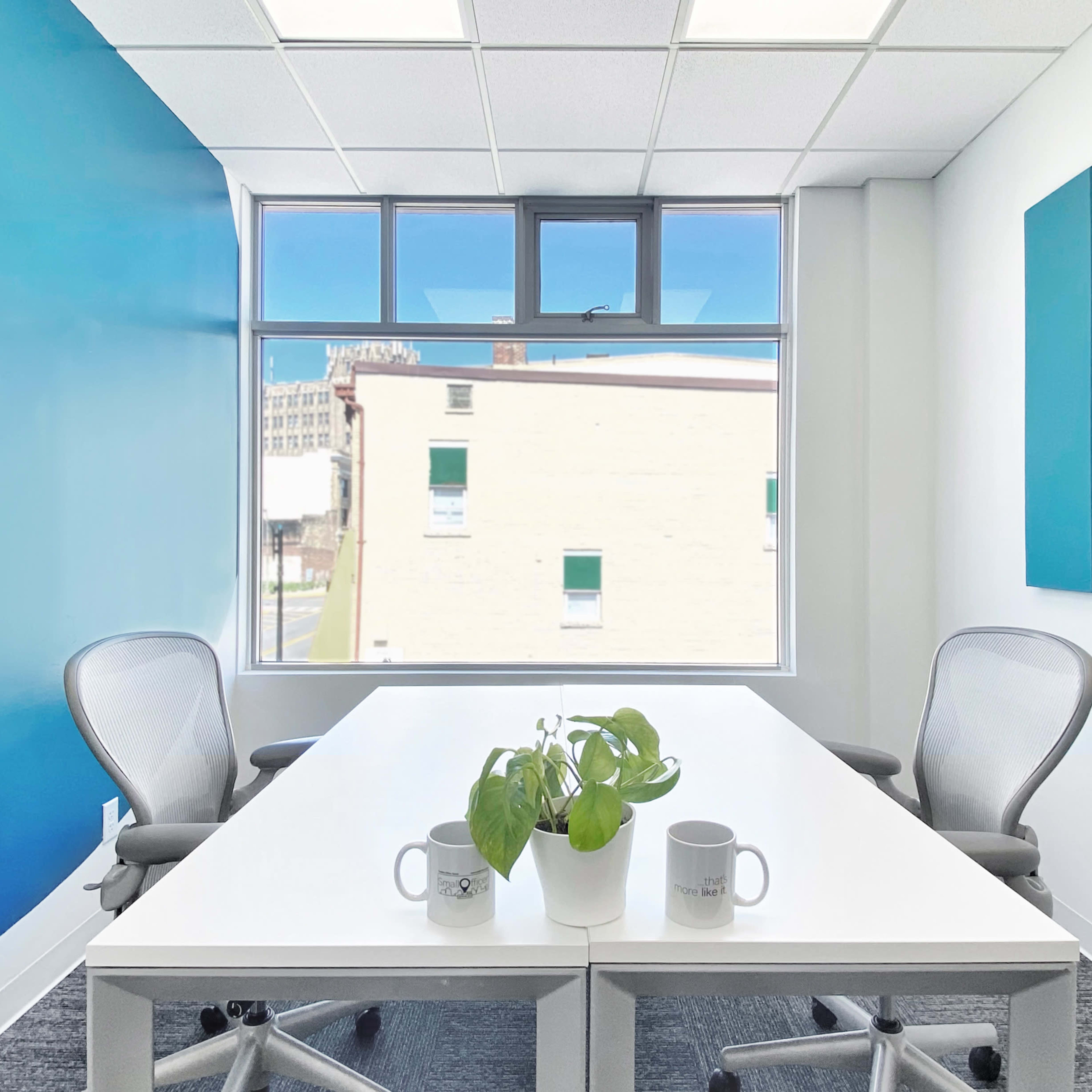 A bright office meeting room features a white table with two chairs, a small plant, and a large window overlooking a city scene.