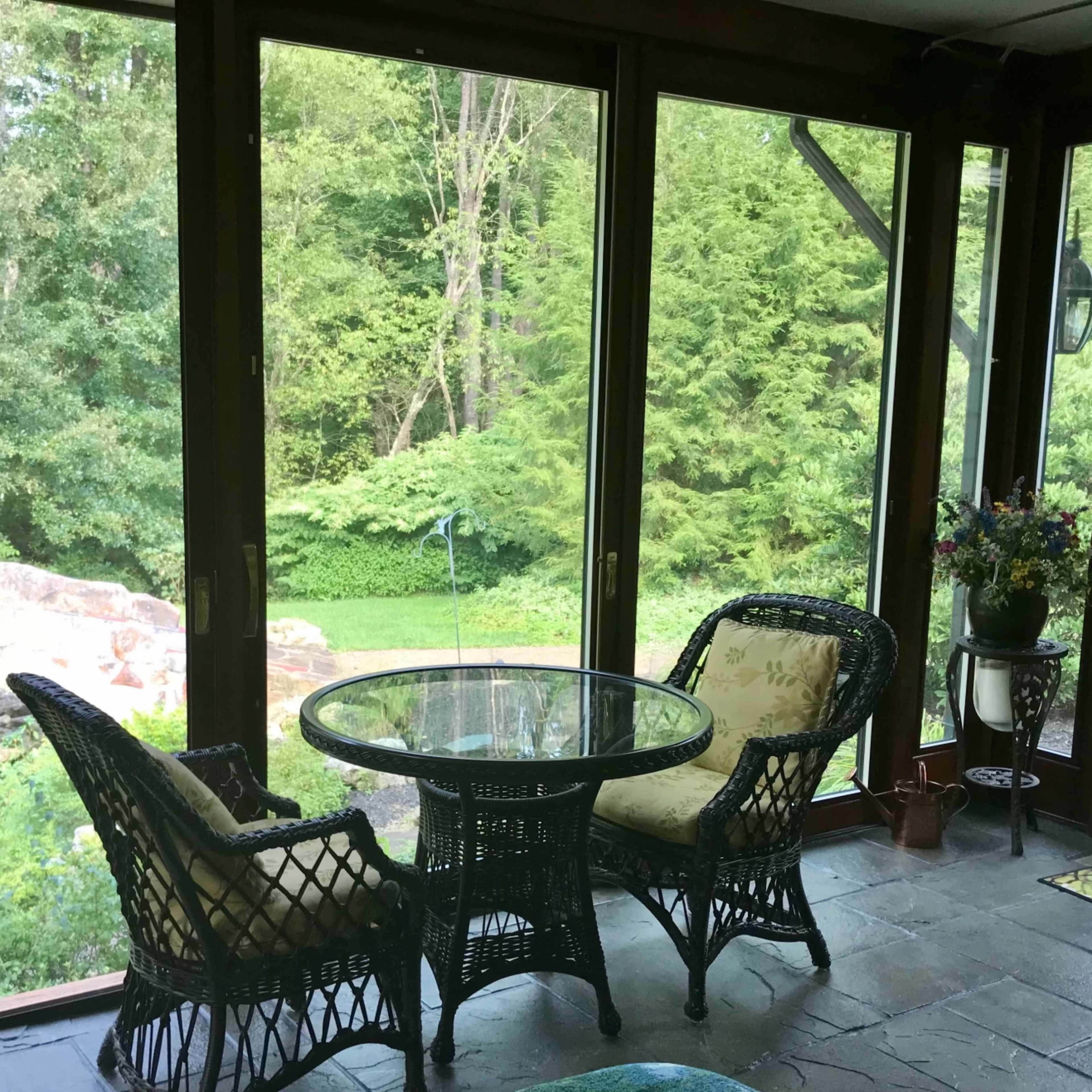 A sunroom features two wicker chairs and a glass table, overlooking a lush garden through large windows.