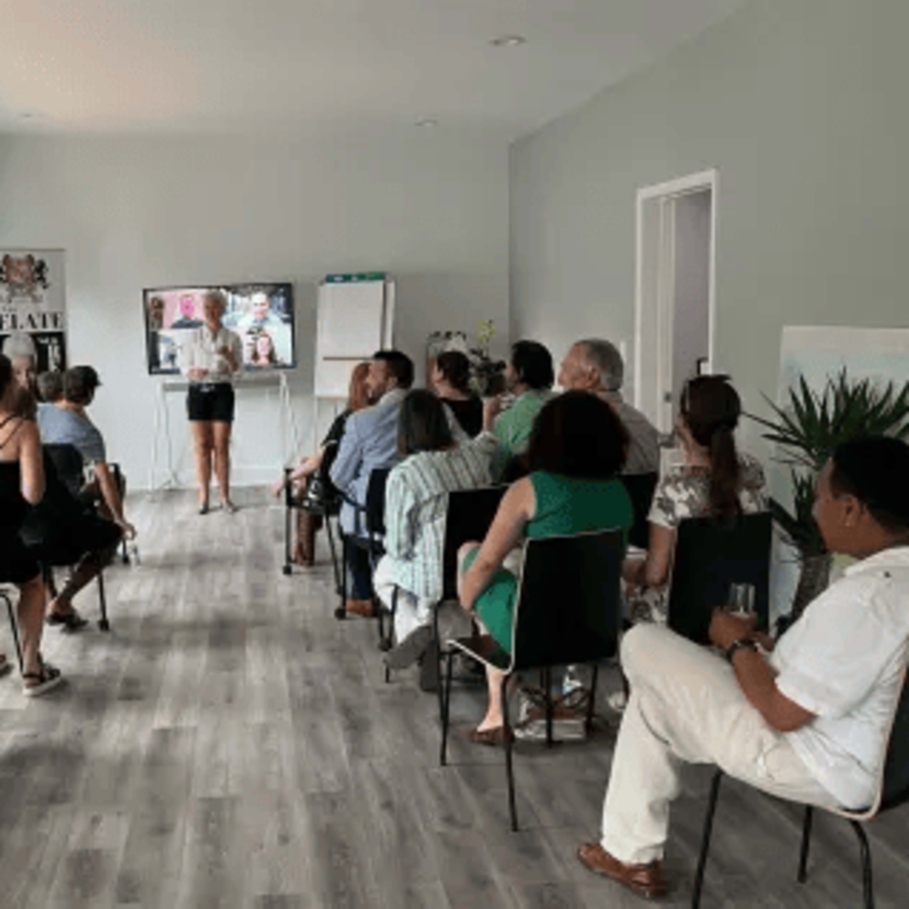 A group of people sits facing a woman presenting in front of a screen in a well-lit room.