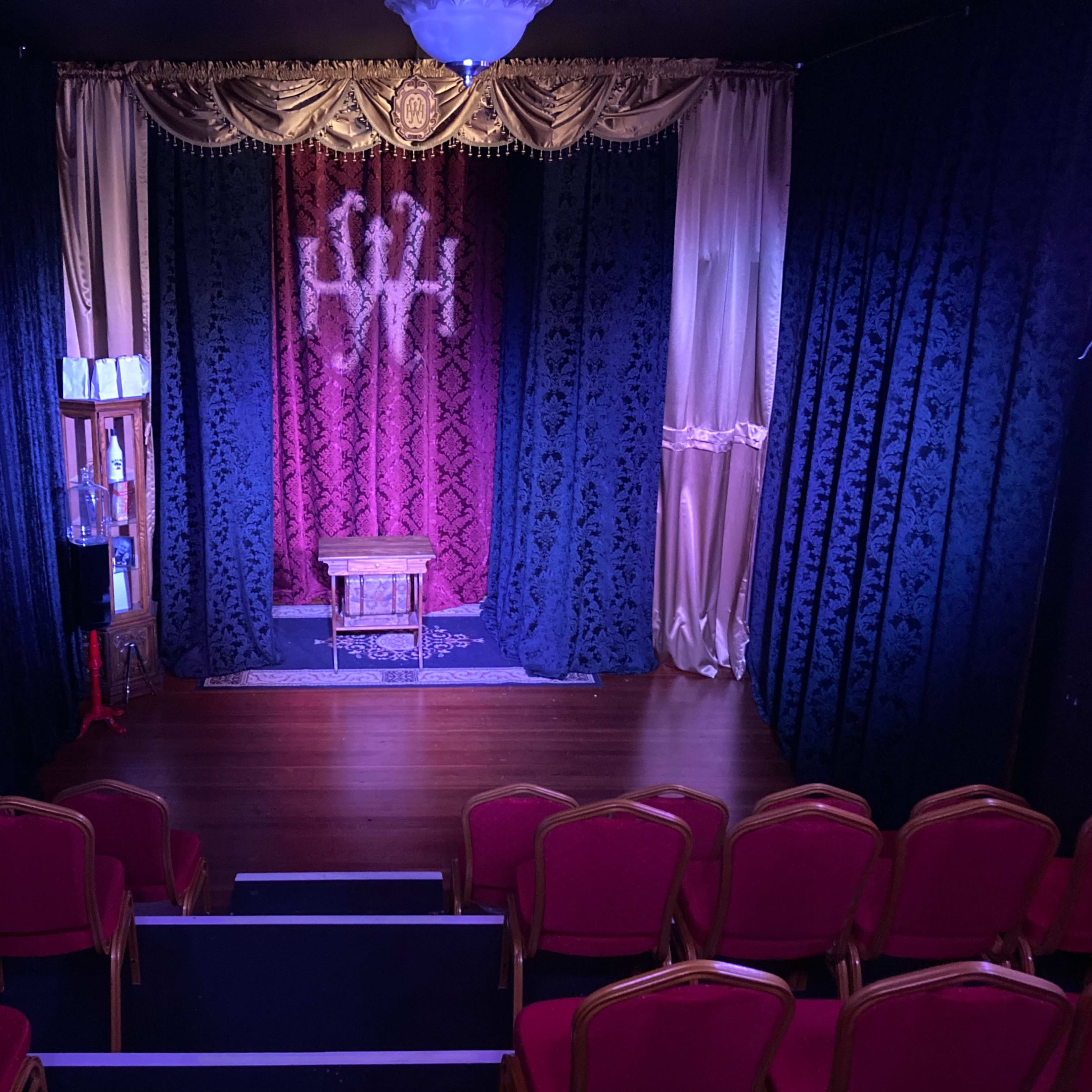 A small theater stage with a decorative backdrop, a wooden table in the center, and red upholstered chairs arranged in front.