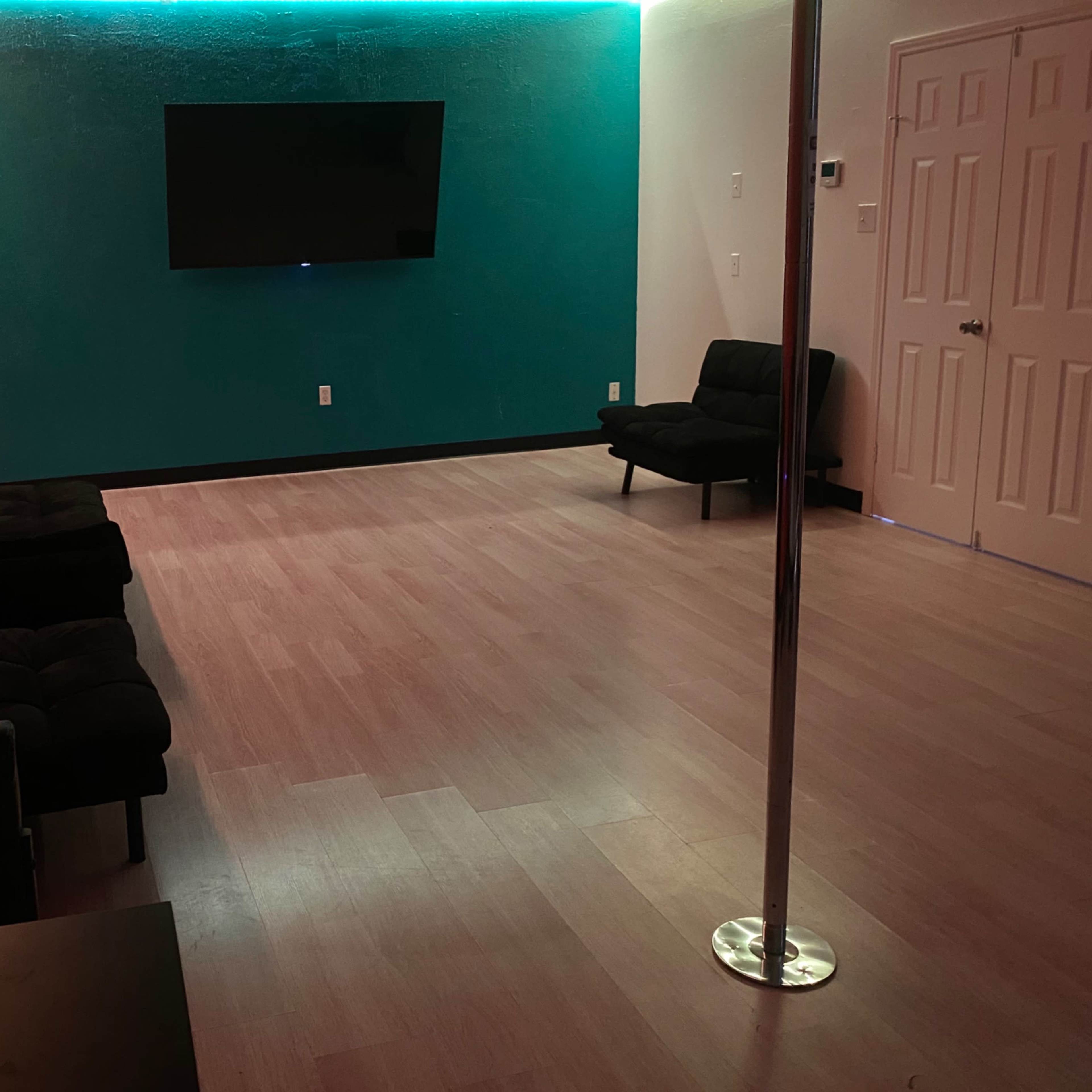 A sparsely furnished room with a television mounted on a teal wall, black couches, a dance pole, and light wooden flooring.