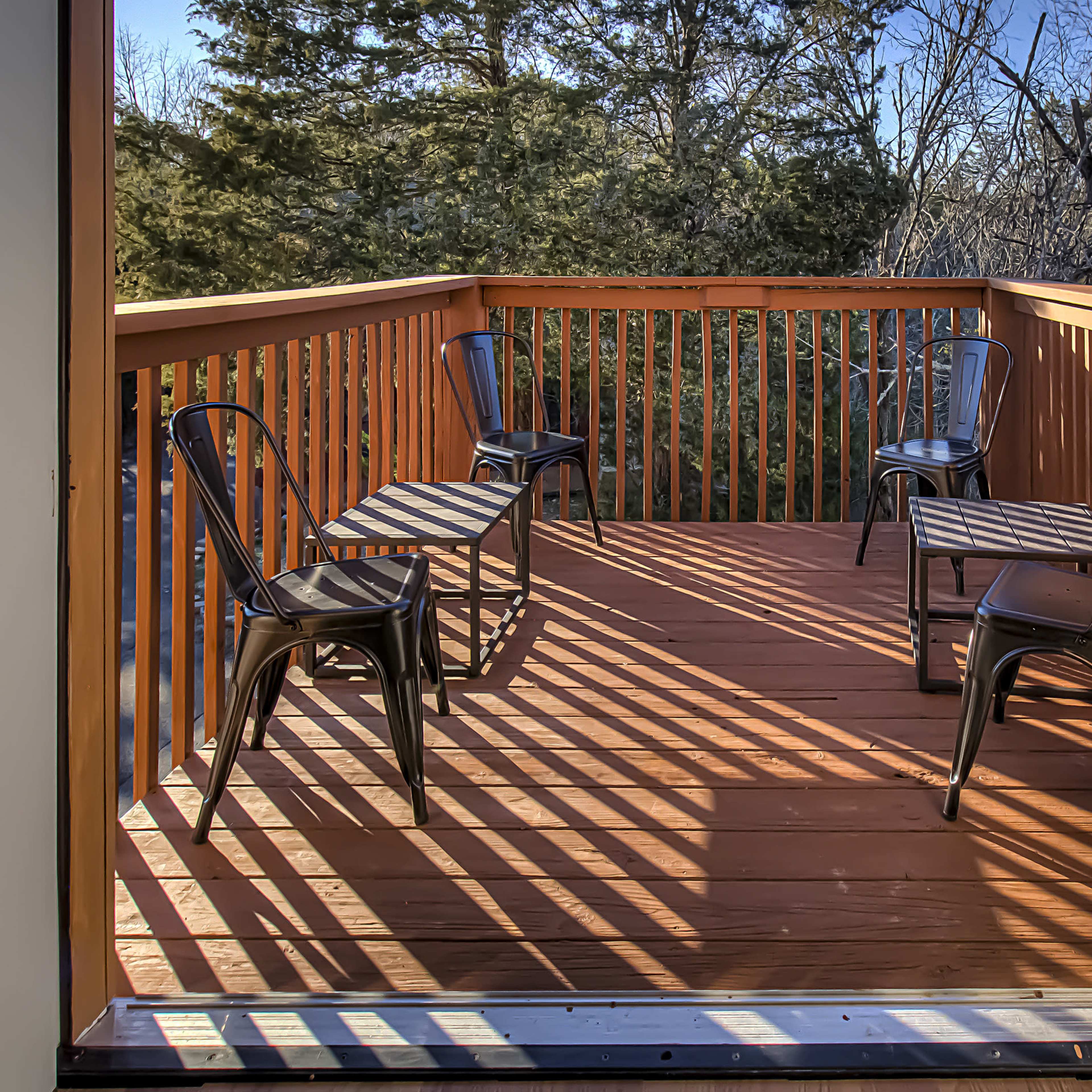 A deck with a wooden railing features four black chairs arranged evenly on the floor, with shadows cast from the railing.