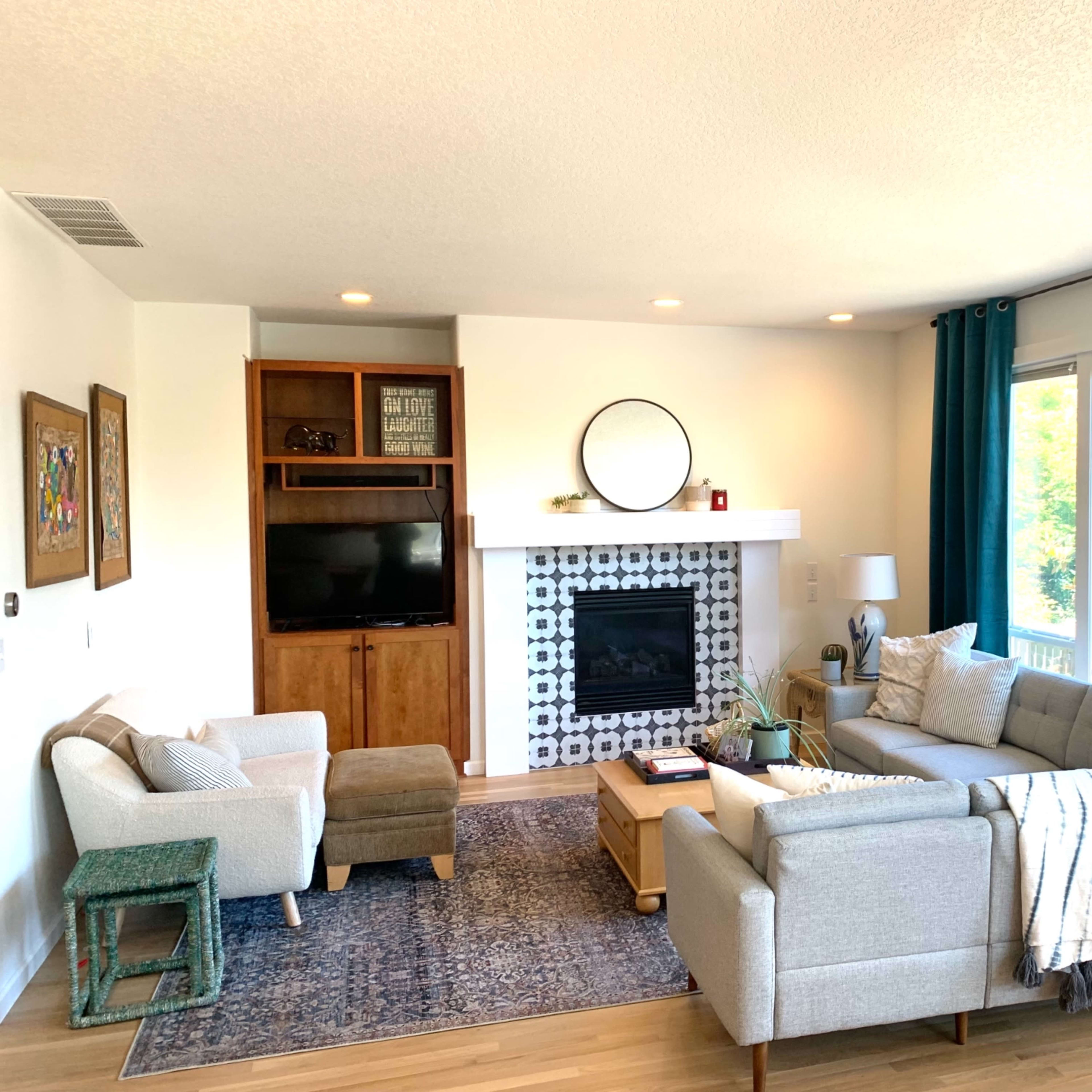 A modern living room features a gray sectional sofa, a wooden coffee table, a patterned area rug, and a fireplace with a circular mirror above it, complemented by large windows and decorative curtains.
