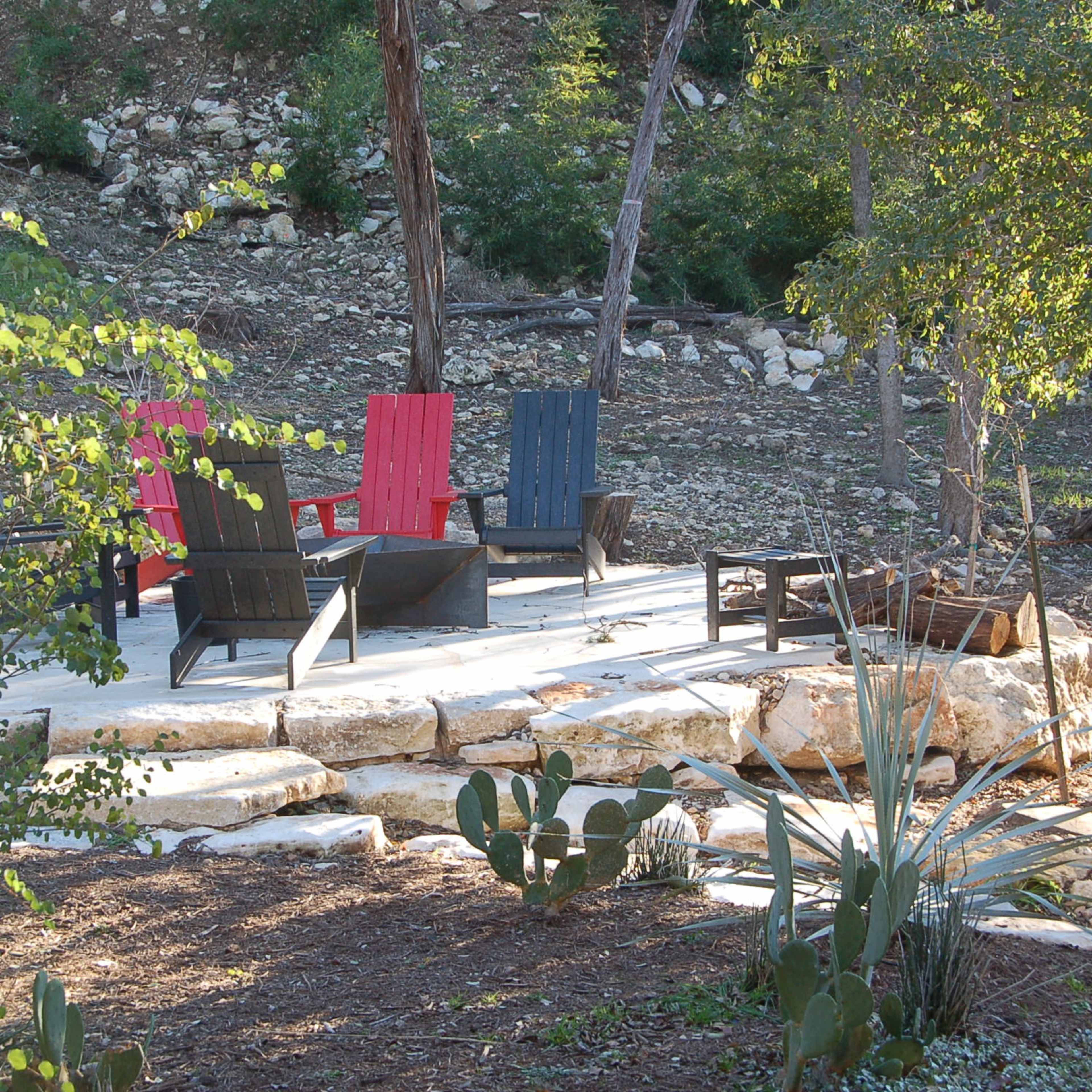 A seating area with four colorful Adirondack chairs arranged around a stone patio in a natural outdoor setting.
