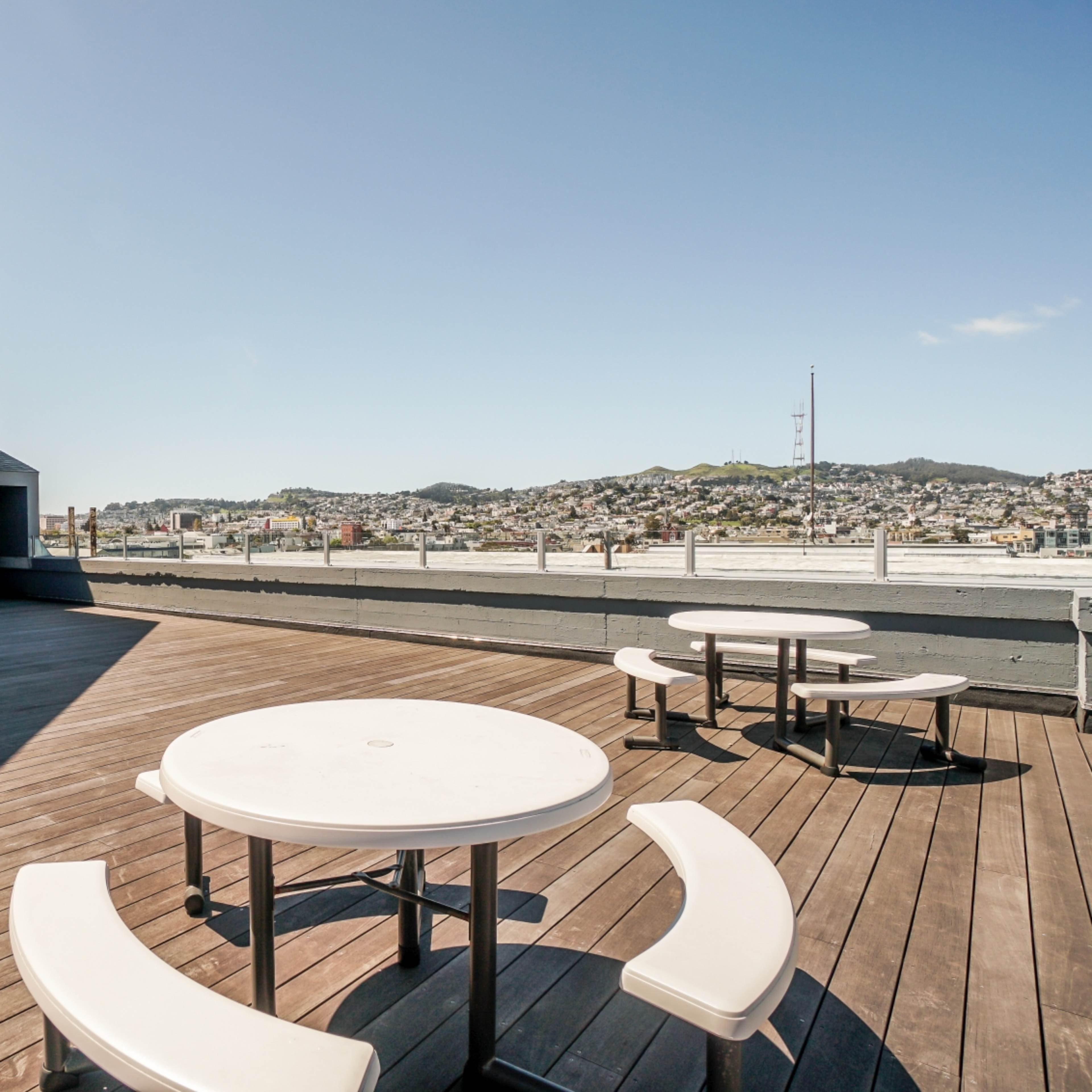 A rooftop terrace with round tables and benches, featuring a view of distant hills and a communication tower against a clear sky.
