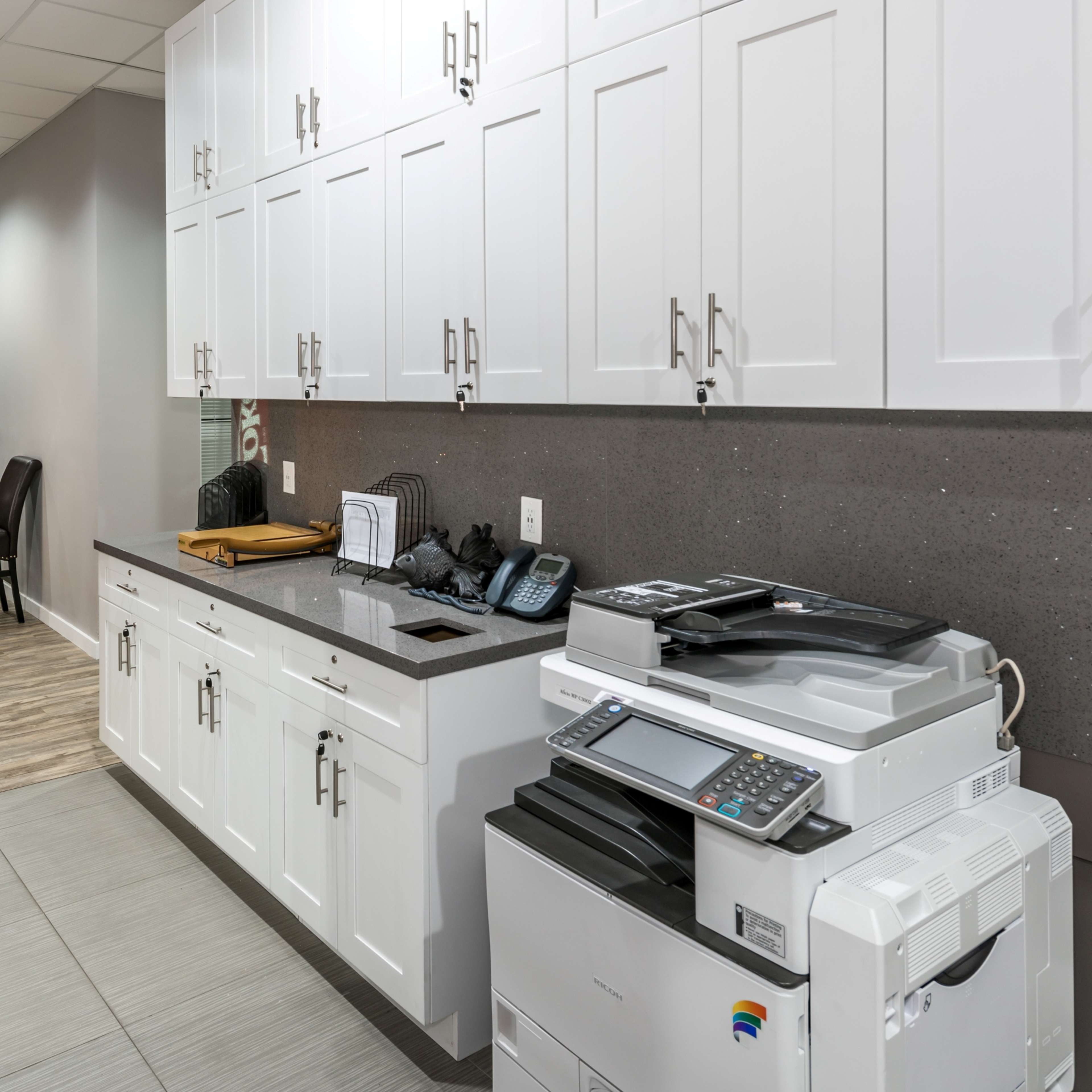 A modern office space with white cabinets, a dark countertop, a printer, and a vending machine in the background.
