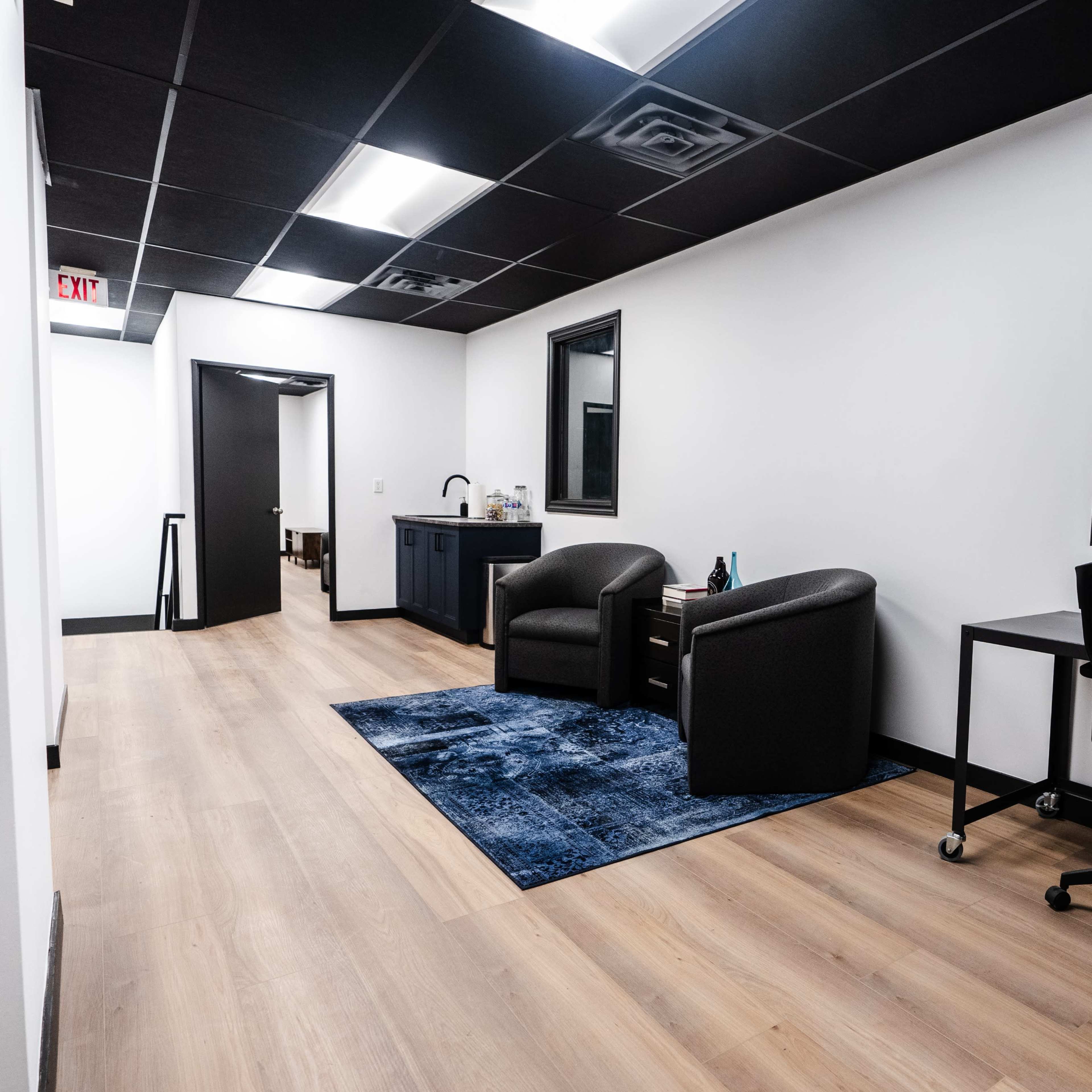 A well-lit office interior featuring a seating area with two chairs, a desk, and a hallway leading to another room.