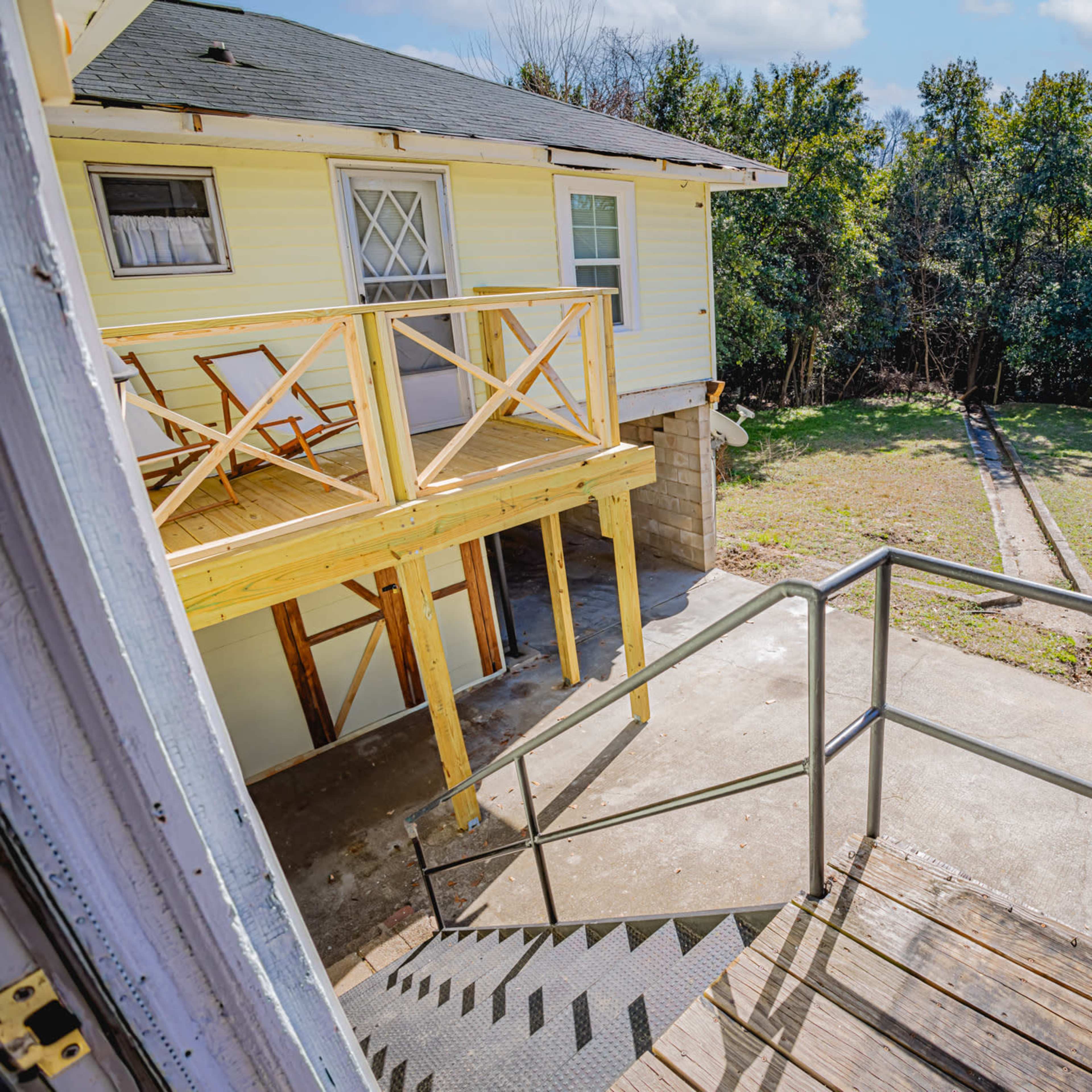 A wooden staircase leads down from a raised deck to a gravel area, adjacent to two separate buildings surrounded by trees.