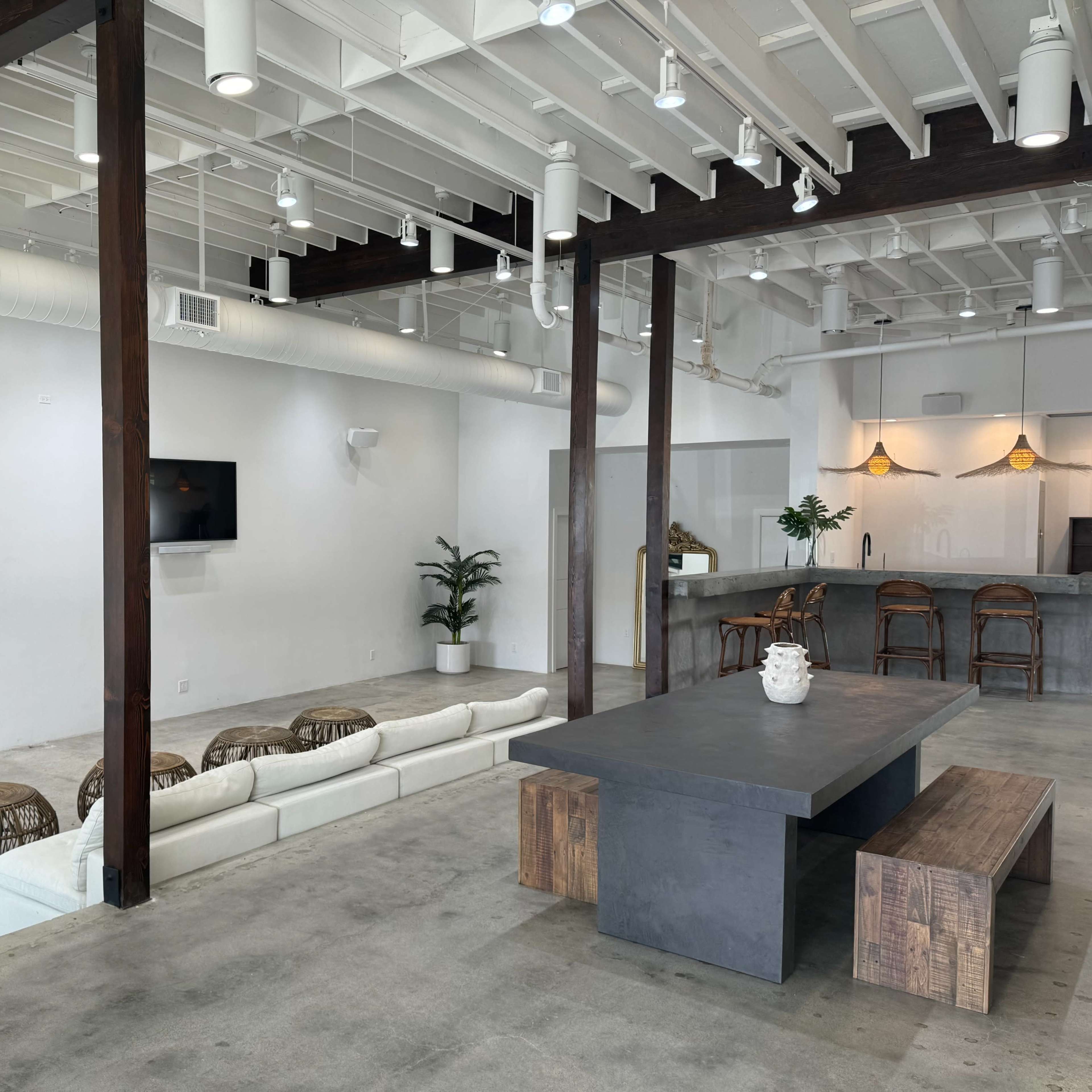 A modern, open-plan lounge area with a long white sofa, a dark wooden table, and barstools, complemented by greenery and minimalistic decor.