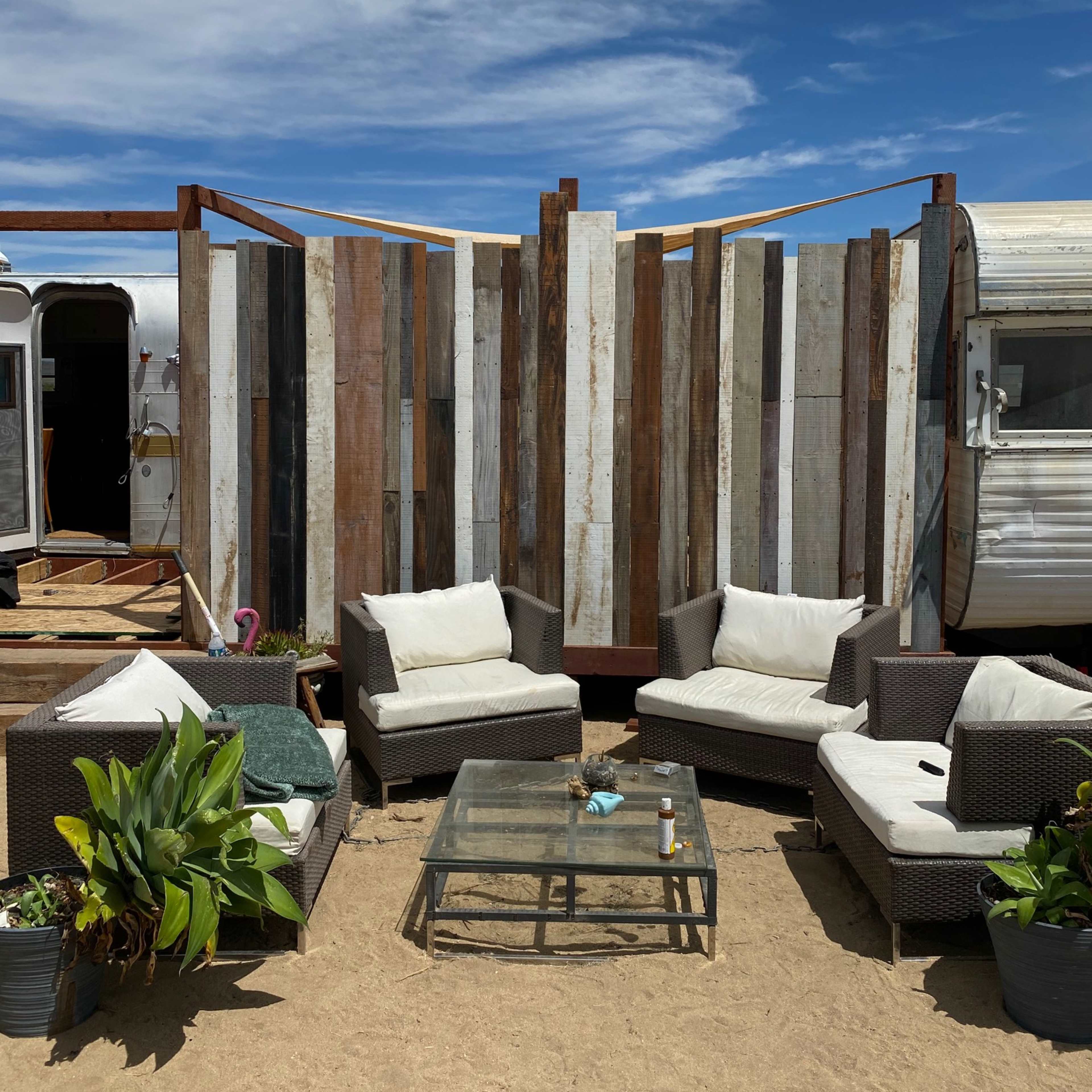 A rustic seating area with wicker furniture and a glass table is set in front of a wooden privacy wall at a sandy location.