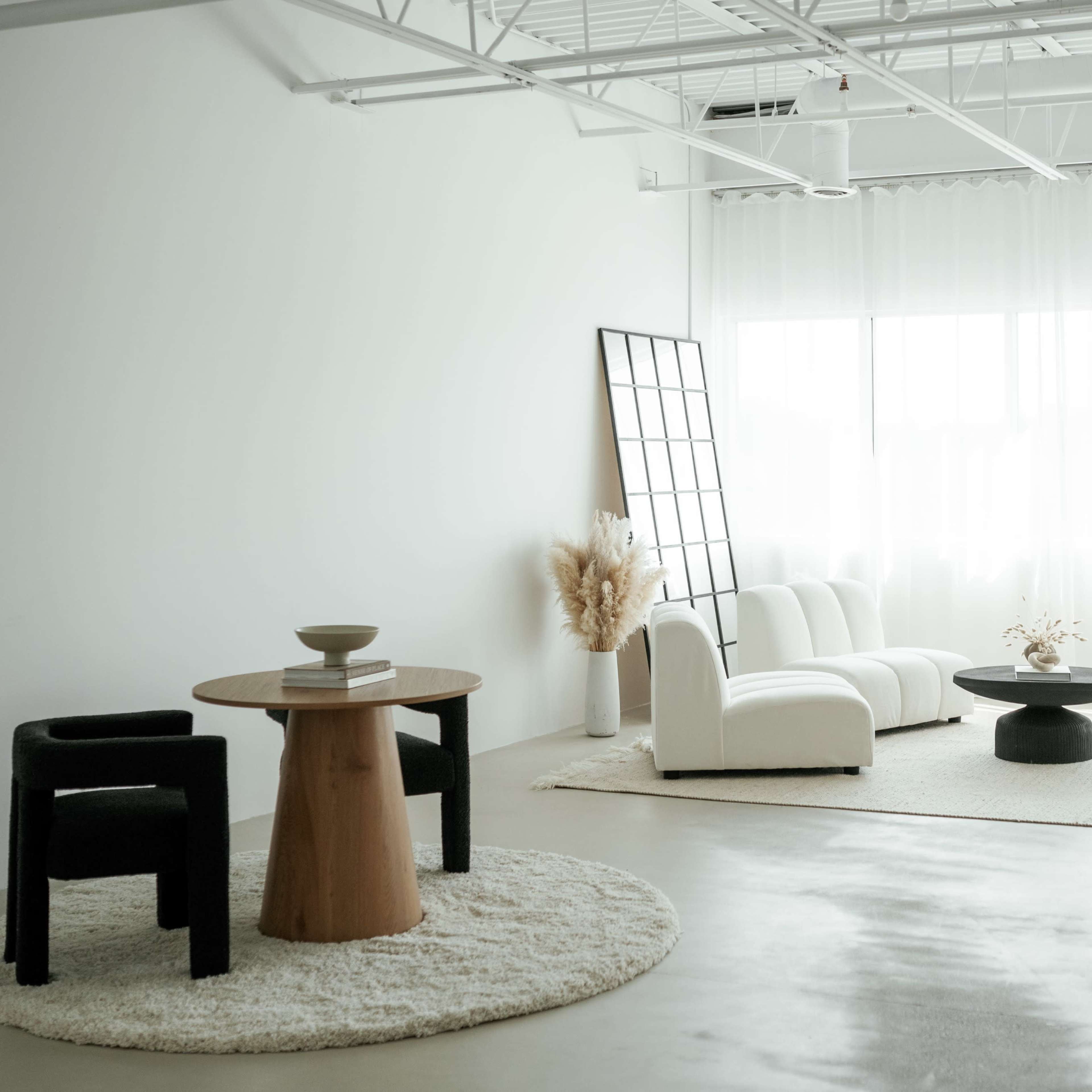 A minimally decorated living room with a white sofa, a round black coffee table, two chairs, and large windows covered by sheer curtains.
