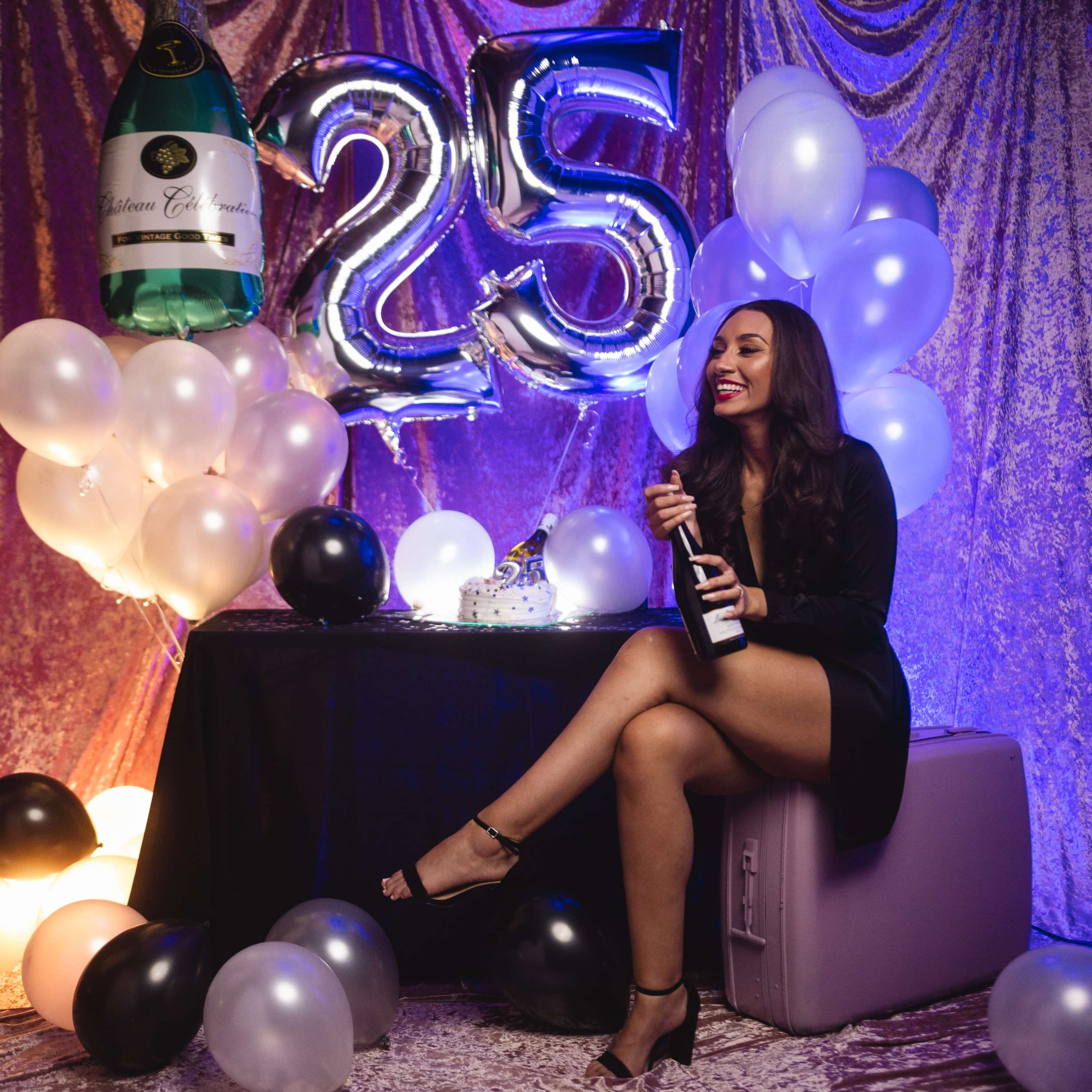 A woman sits on a box holding a bottle of champagne, surrounded by balloons and a large "25" balloon, with a cake on a table nearby.