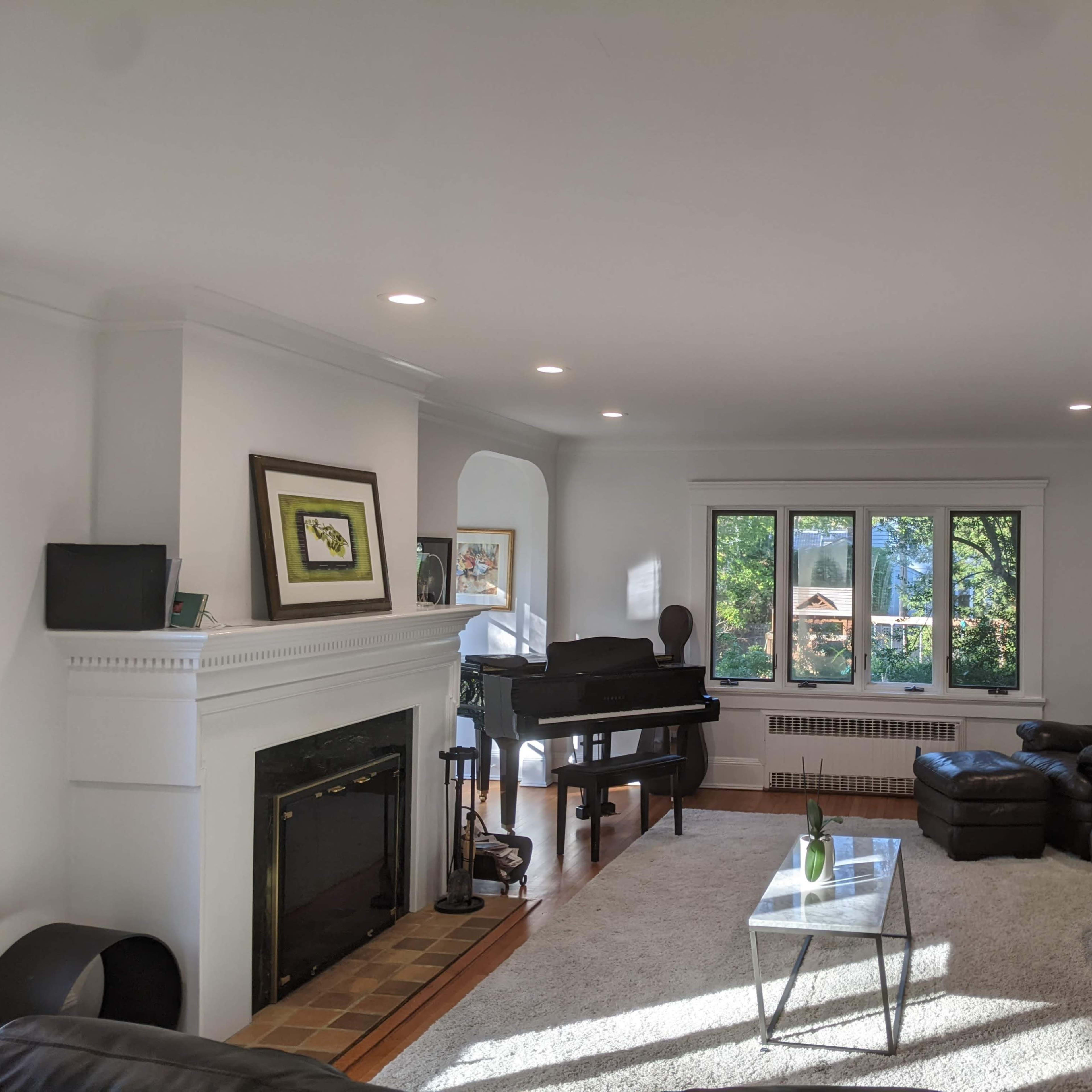 A spacious living room with dark leather sofas, a grand piano, a fireplace, and large windows overlooking greenery.