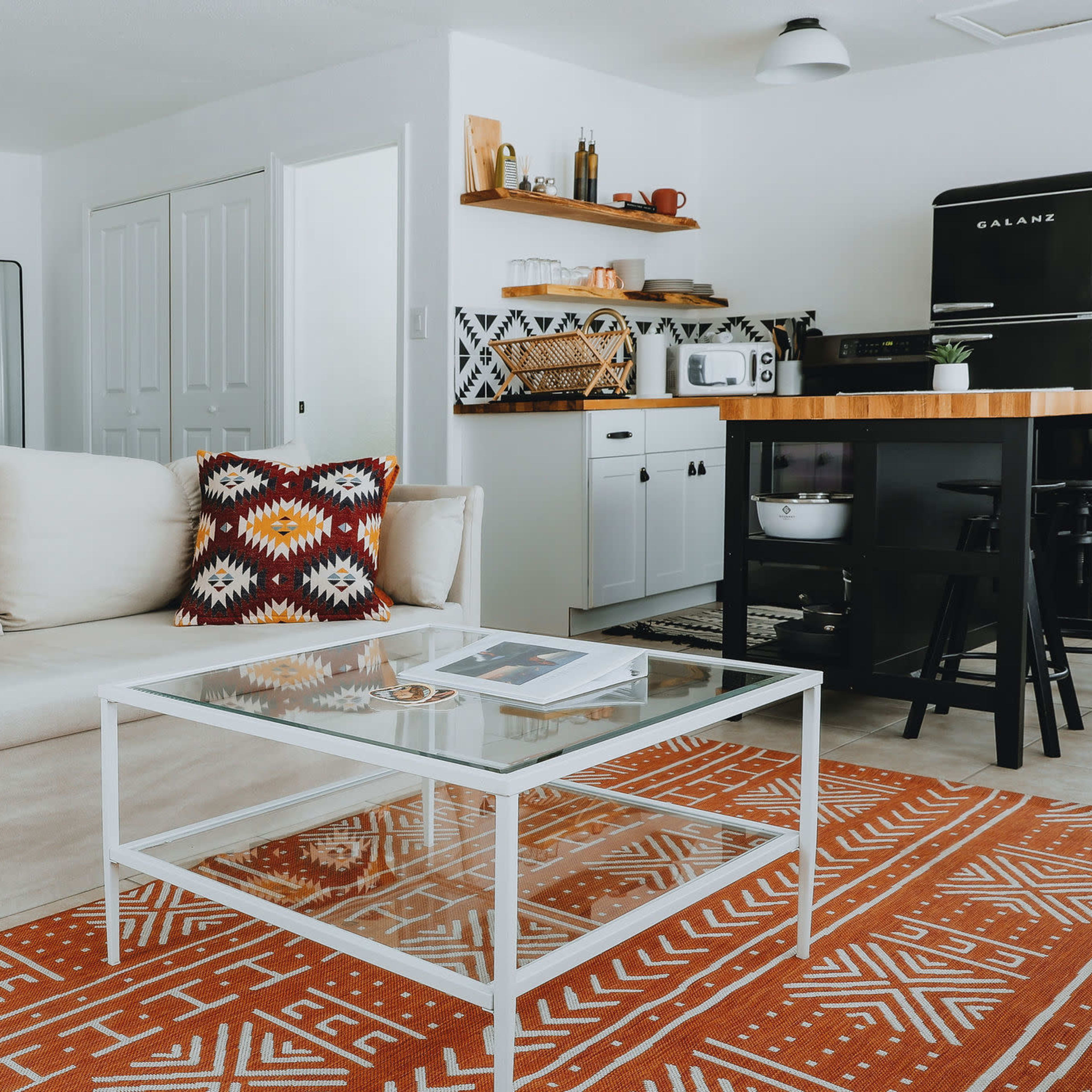 A modern living room with a white couch, a glass coffee table, a patterned rug, and a kitchen area in the background with black and white appliances.