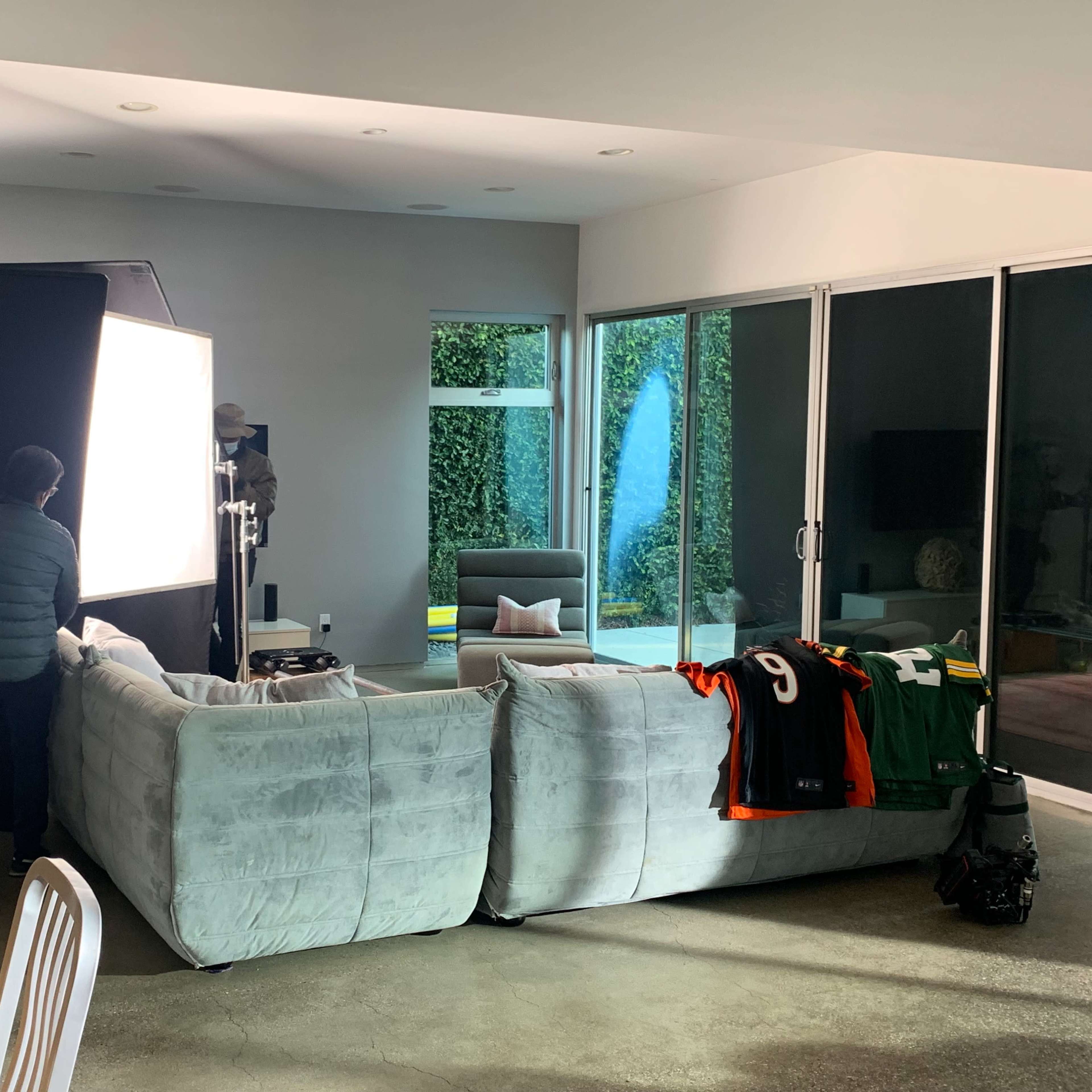 A person adjusts a lighting setup in a modern living room featuring a sectional sofa and a window view of greenery.