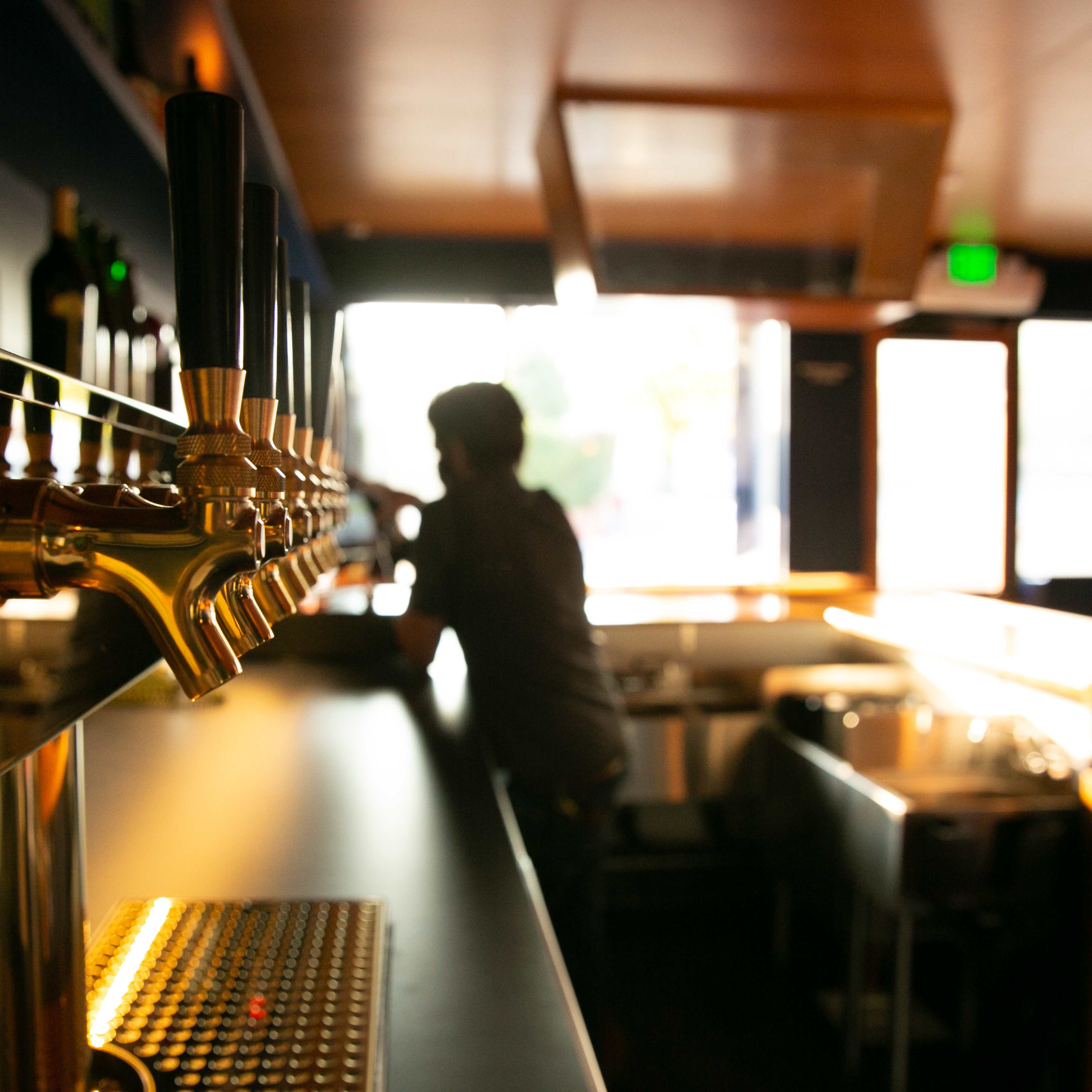 A silhouette of a person stands near a bar with several beer taps in a dimly lit space.