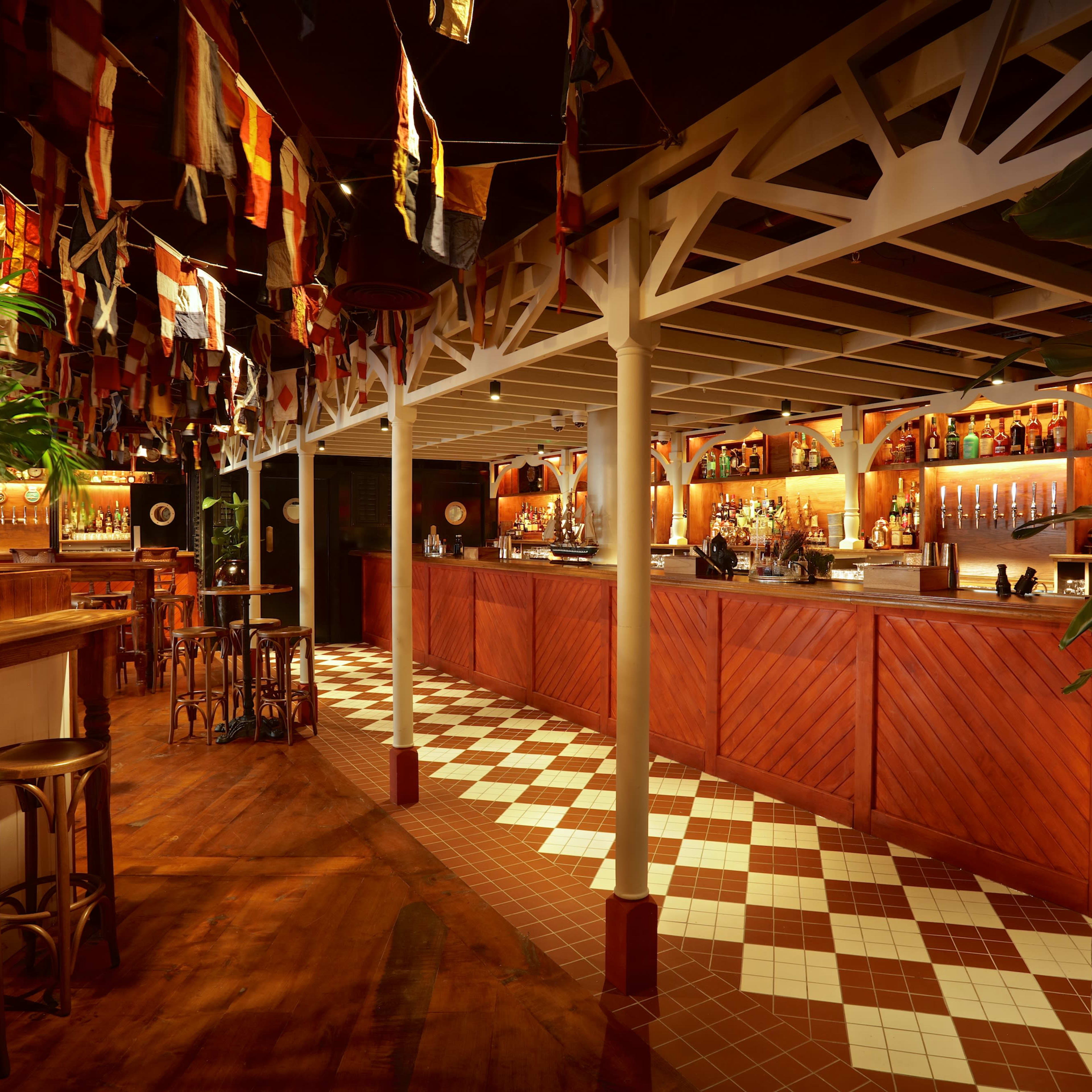 A warmly lit bar interior with wooden furnishings, a long counter stocked with bottles, and colorful flags hanging from the ceiling.