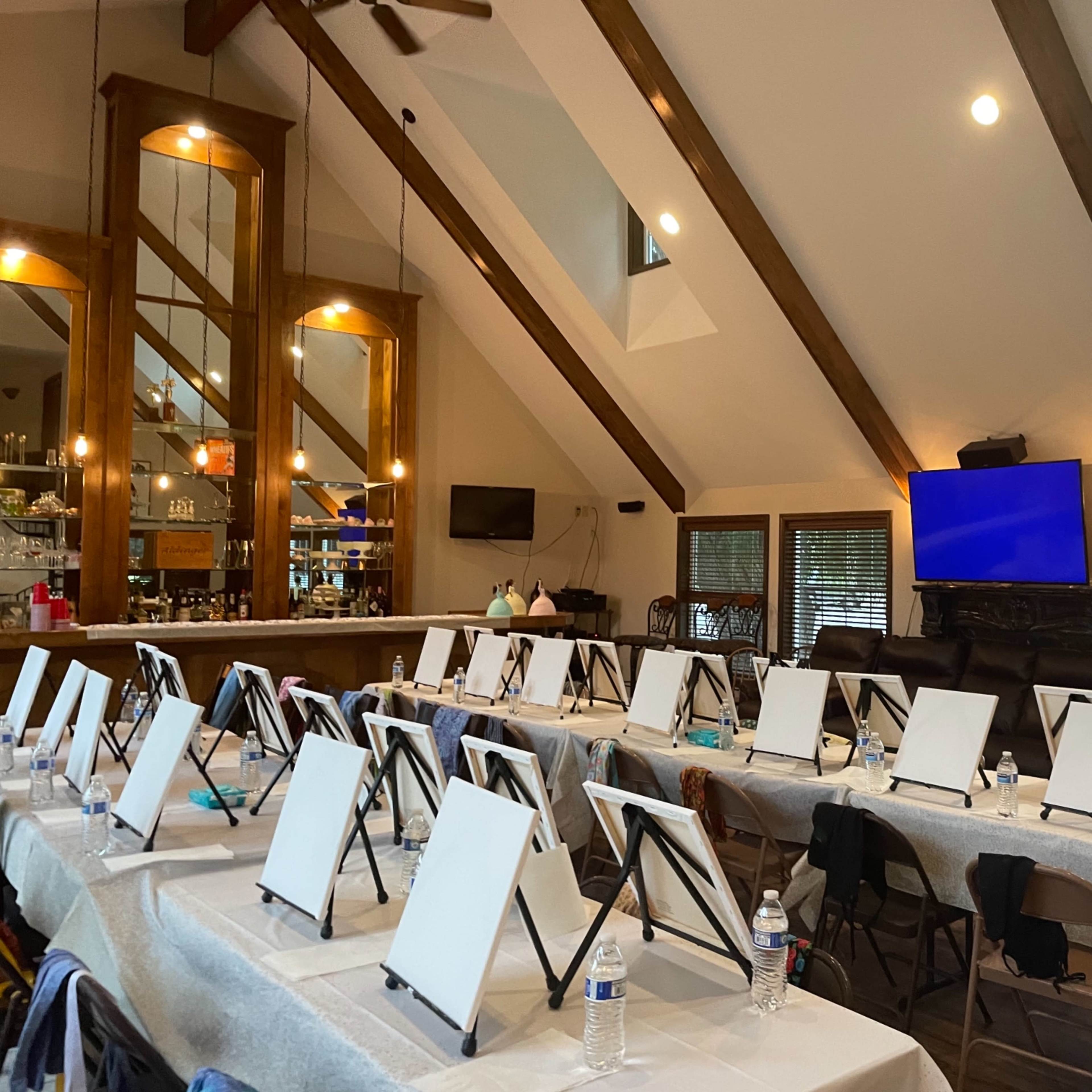 An indoor space set up for a painting class, featuring multiple empty easels arranged neatly on tables with water bottles and a bar area visible in the background.