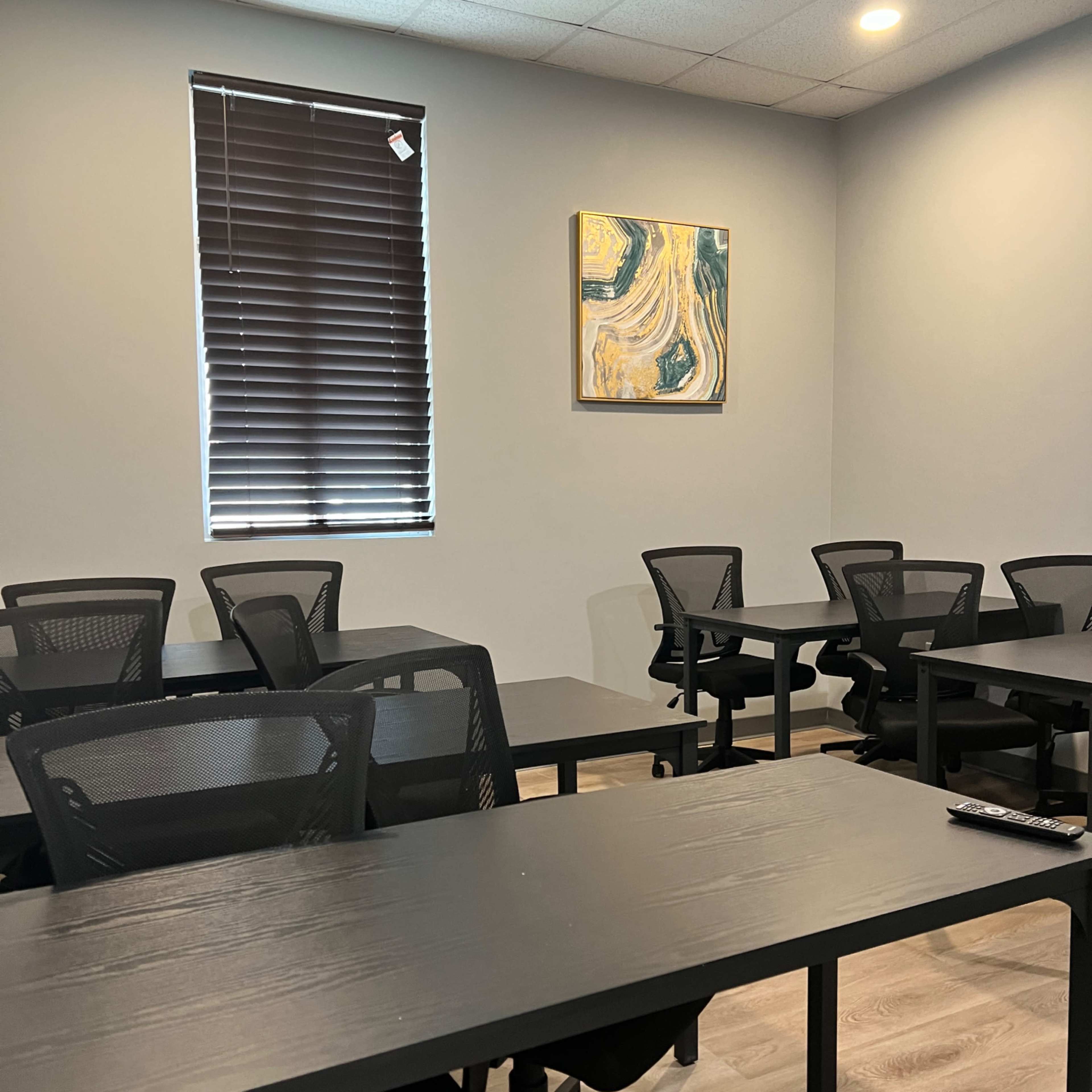 A classroom features several black tables and chairs arranged neatly, with a large abstract painting hanging on the wall.