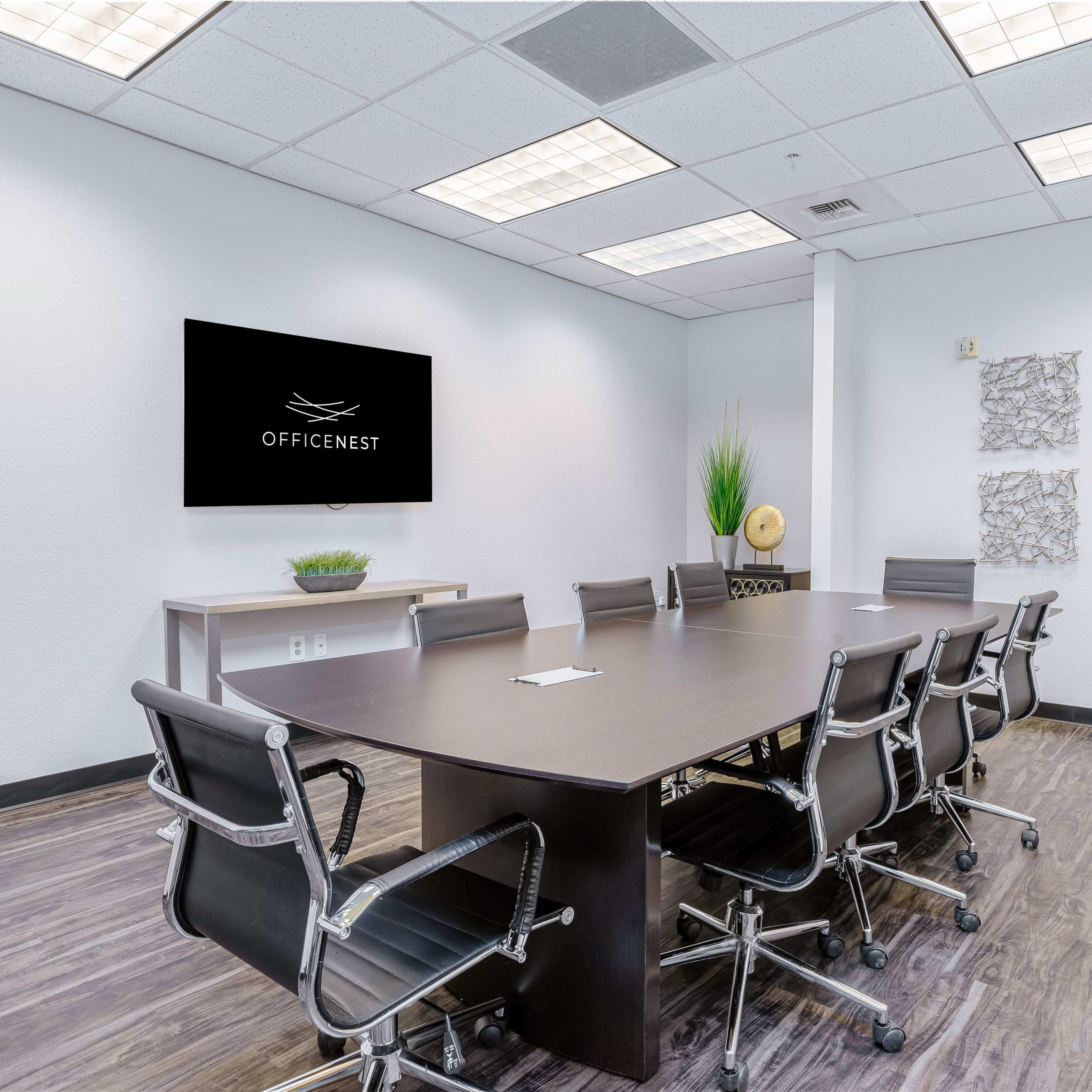 A modern conference room features a long table surrounded by ergonomic chairs, with a wall-mounted TV and decorative wall art.