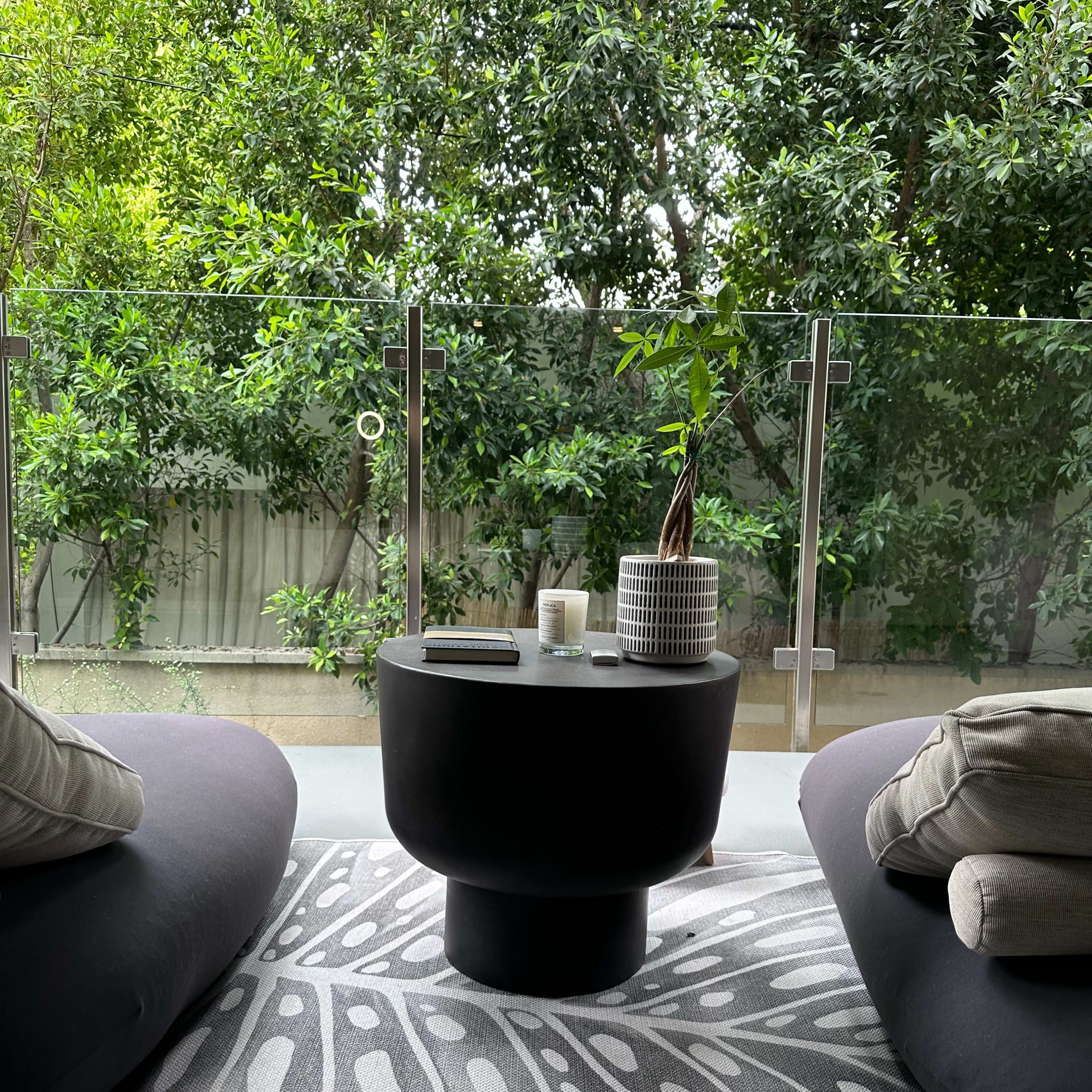A balcony features two rounded chairs, a cylindrical table, and a rug, all set against a backdrop of lush greenery.