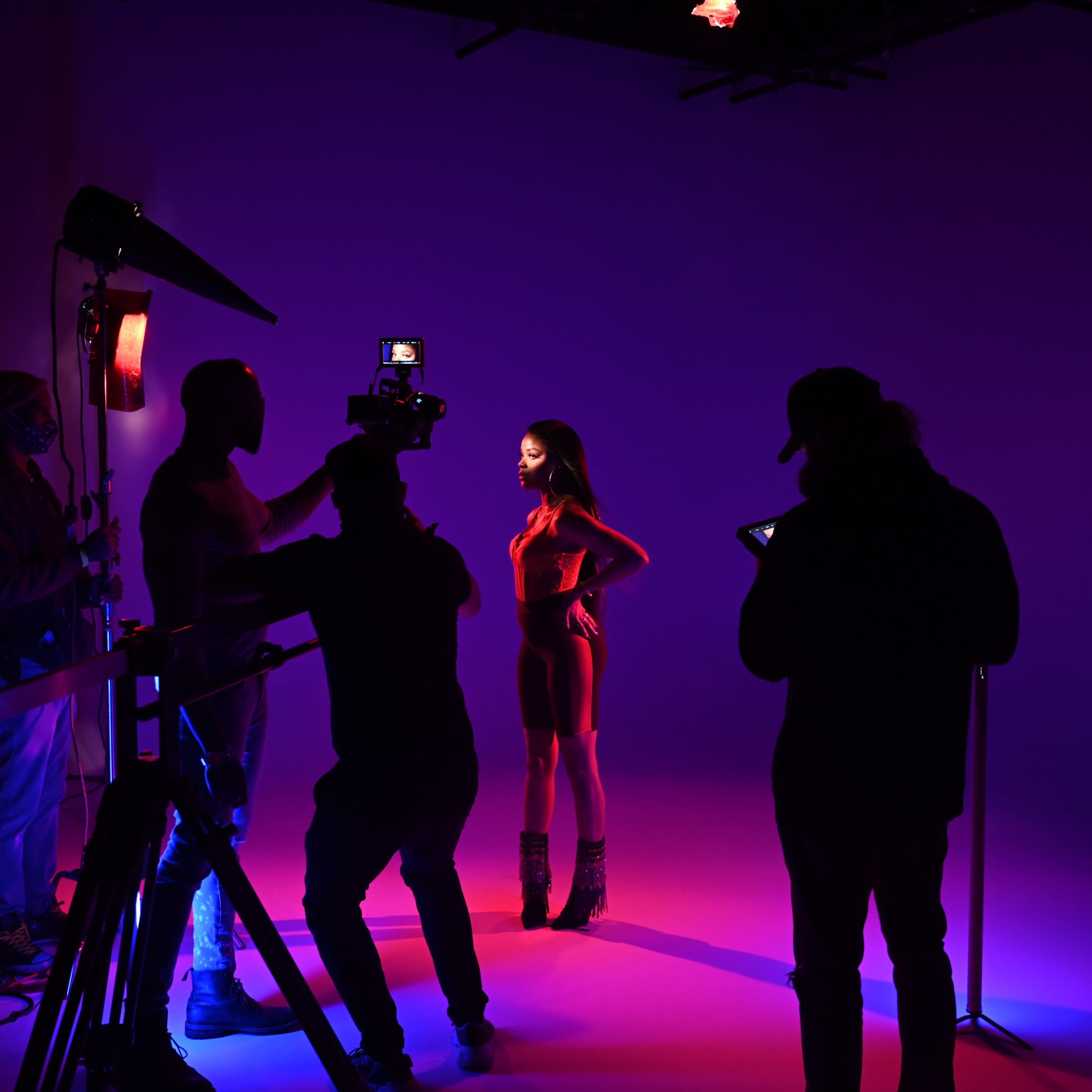 A model poses against a brightly lit backdrop while a team of crew members operates cameras and lighting equipment around her.