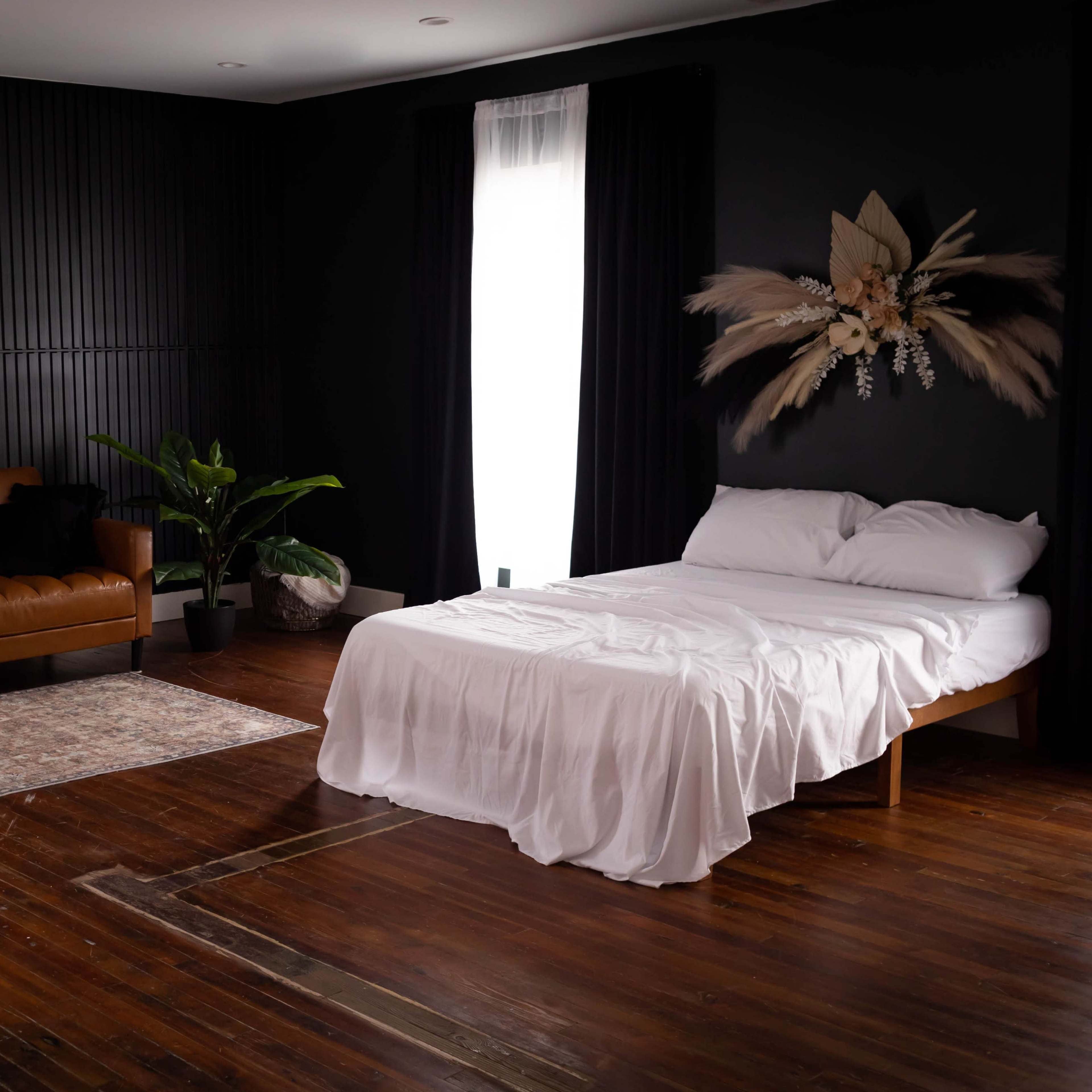 A dimly lit room featuring a brown leather sofa, a white bed with a plain cover, a patterned rug, and several indoor plants.
