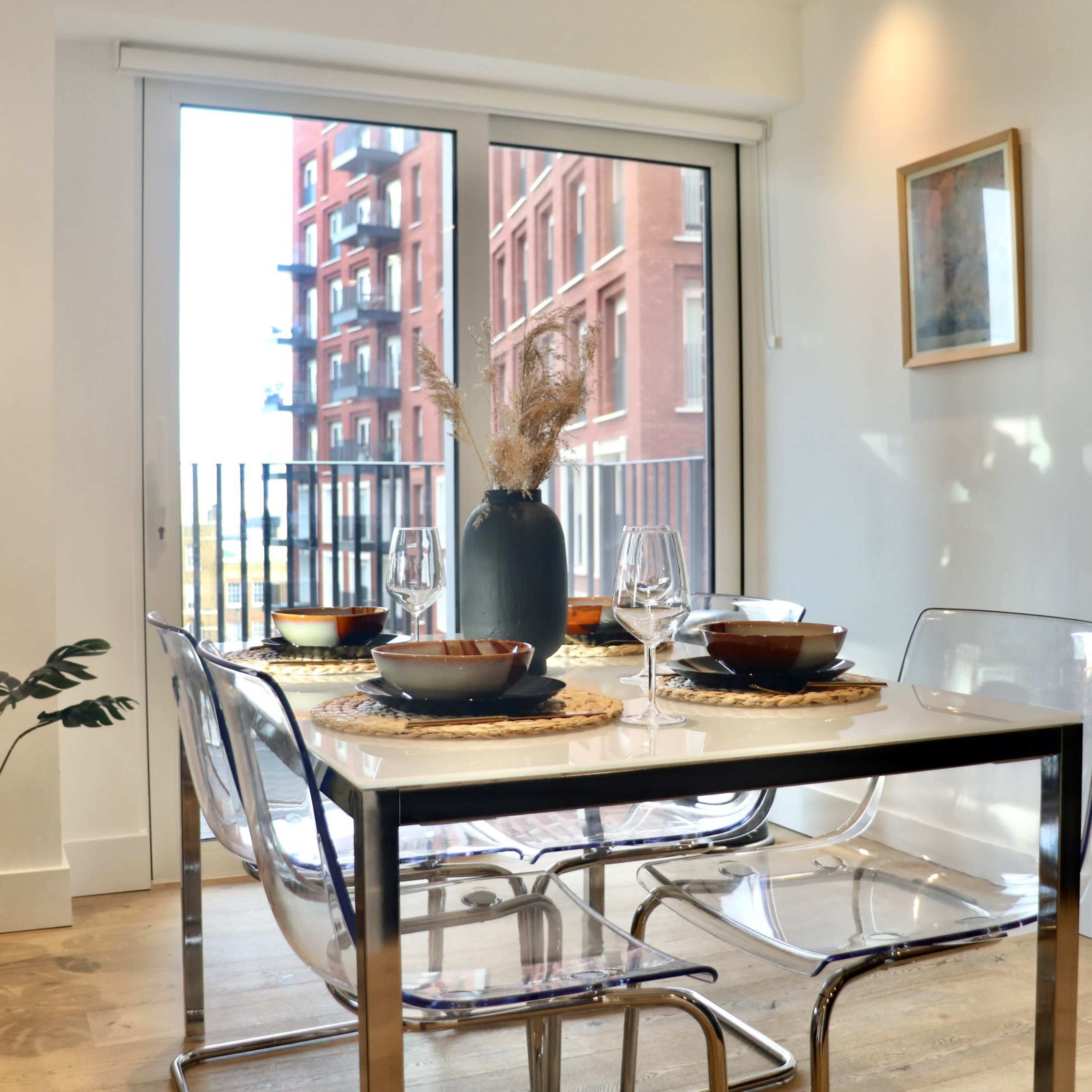 A dining table set for four is arranged next to a large window overlooking a building with red brick exterior.