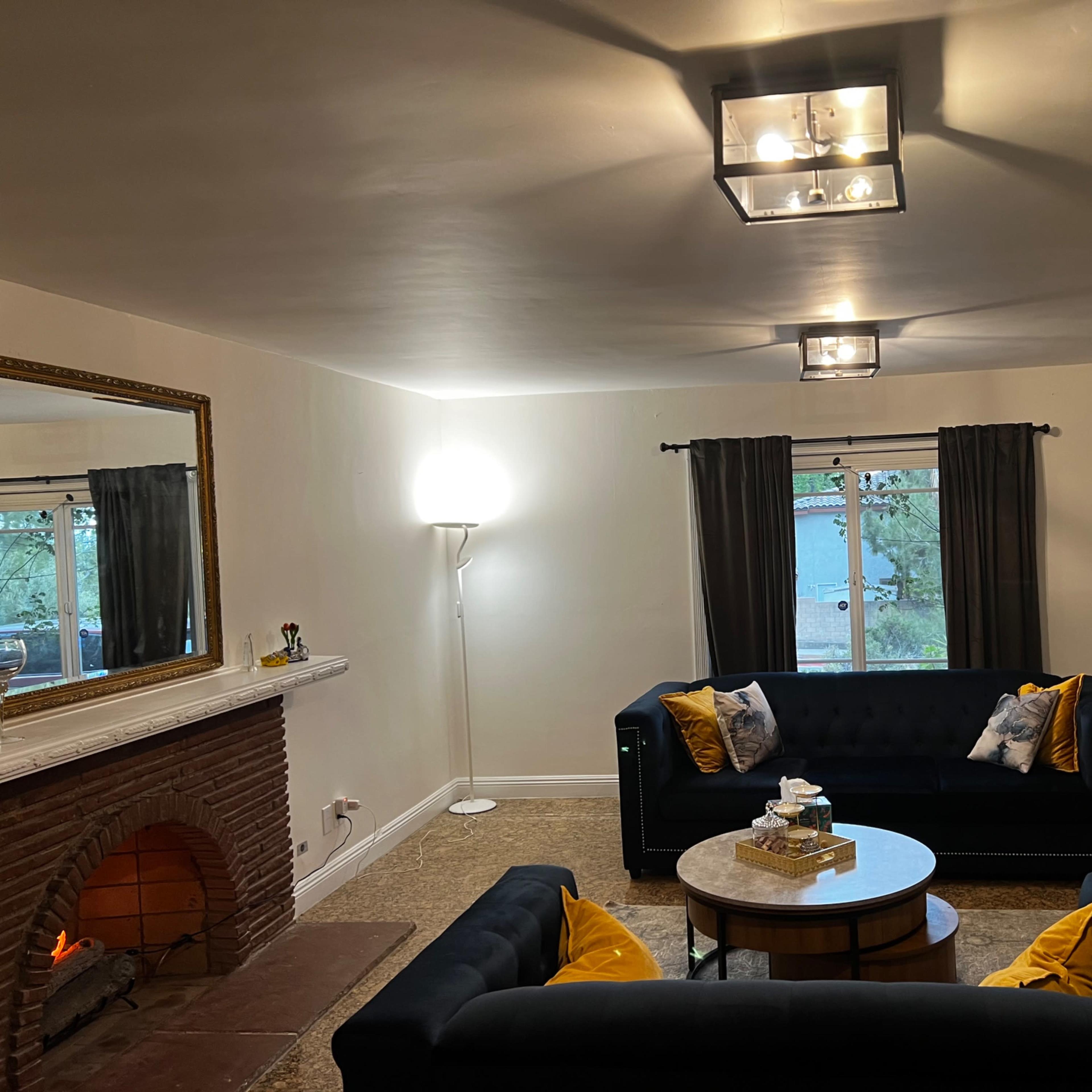 A living room features two black sofas adorned with yellow pillows, a round coffee table in the center, and a fireplace against one wall, with a lamp and windows providing natural light.