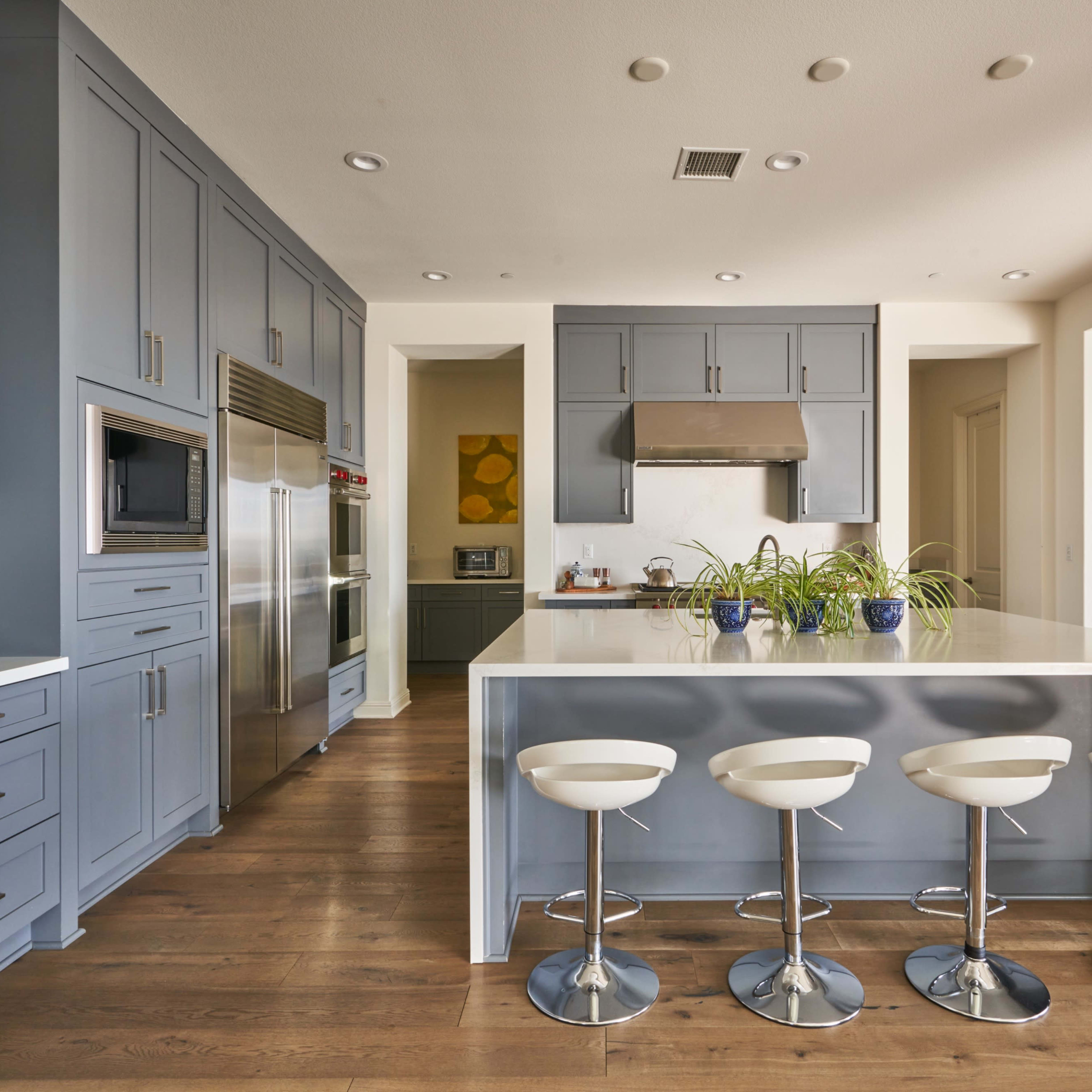A modern kitchen features blue cabinetry, a central island with four white bar stools, and a pantry filled with colorful food items.