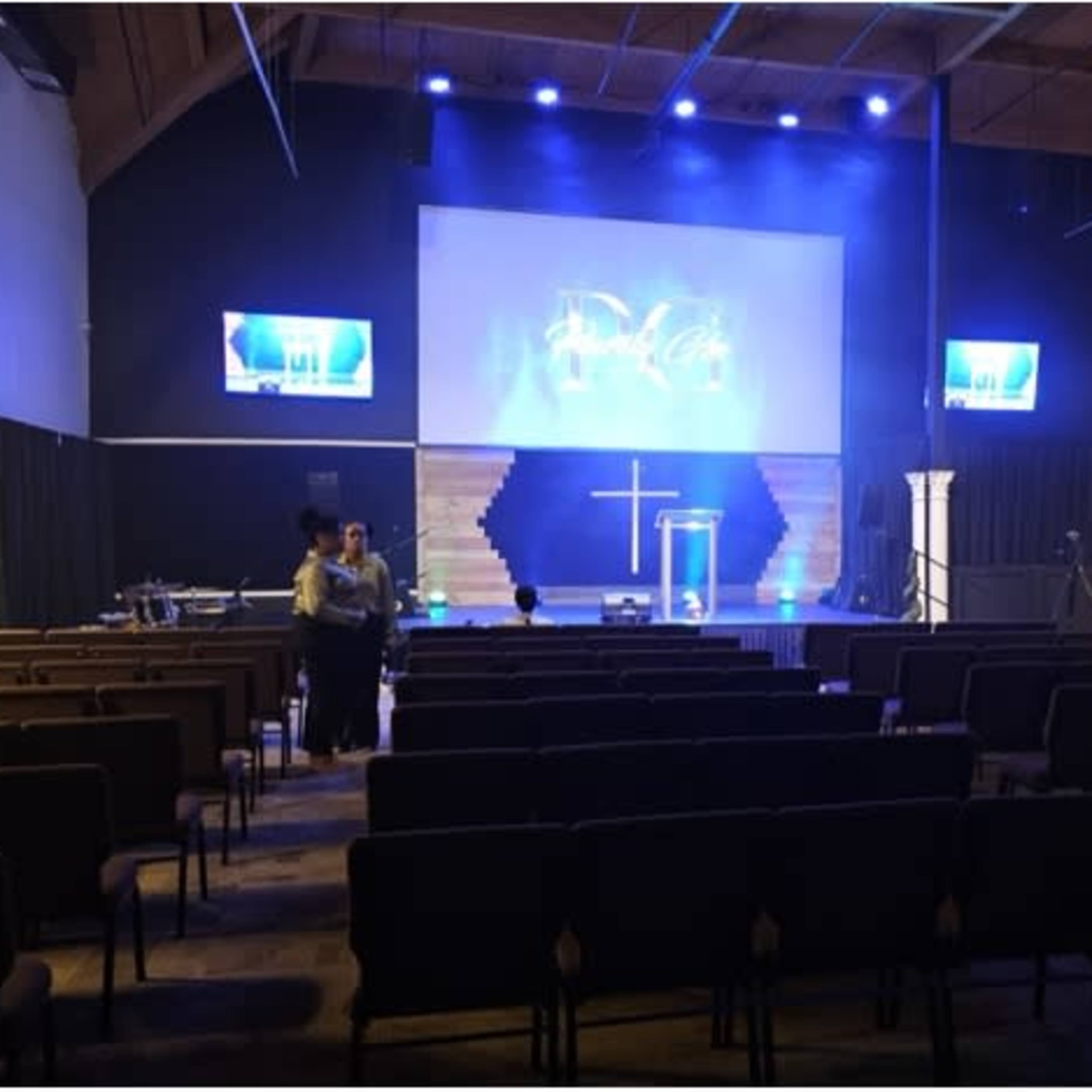 The interior of a church is shown, featuring rows of empty chairs facing a stage with a cross and illuminated screens.