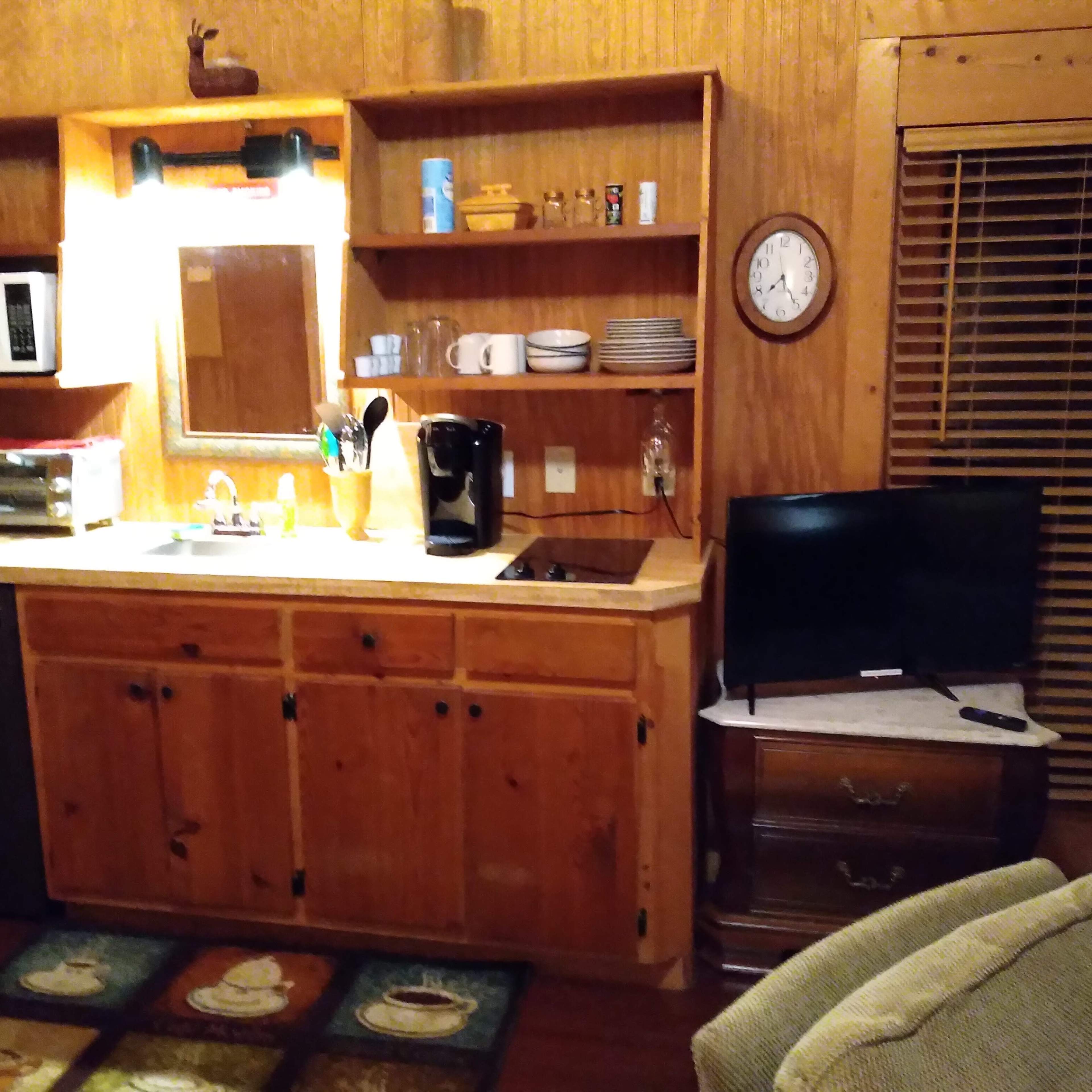 A wooden kitchen area with a microwave, coffee maker, sink, and a small television, along with shelves displaying dishes and utensils.