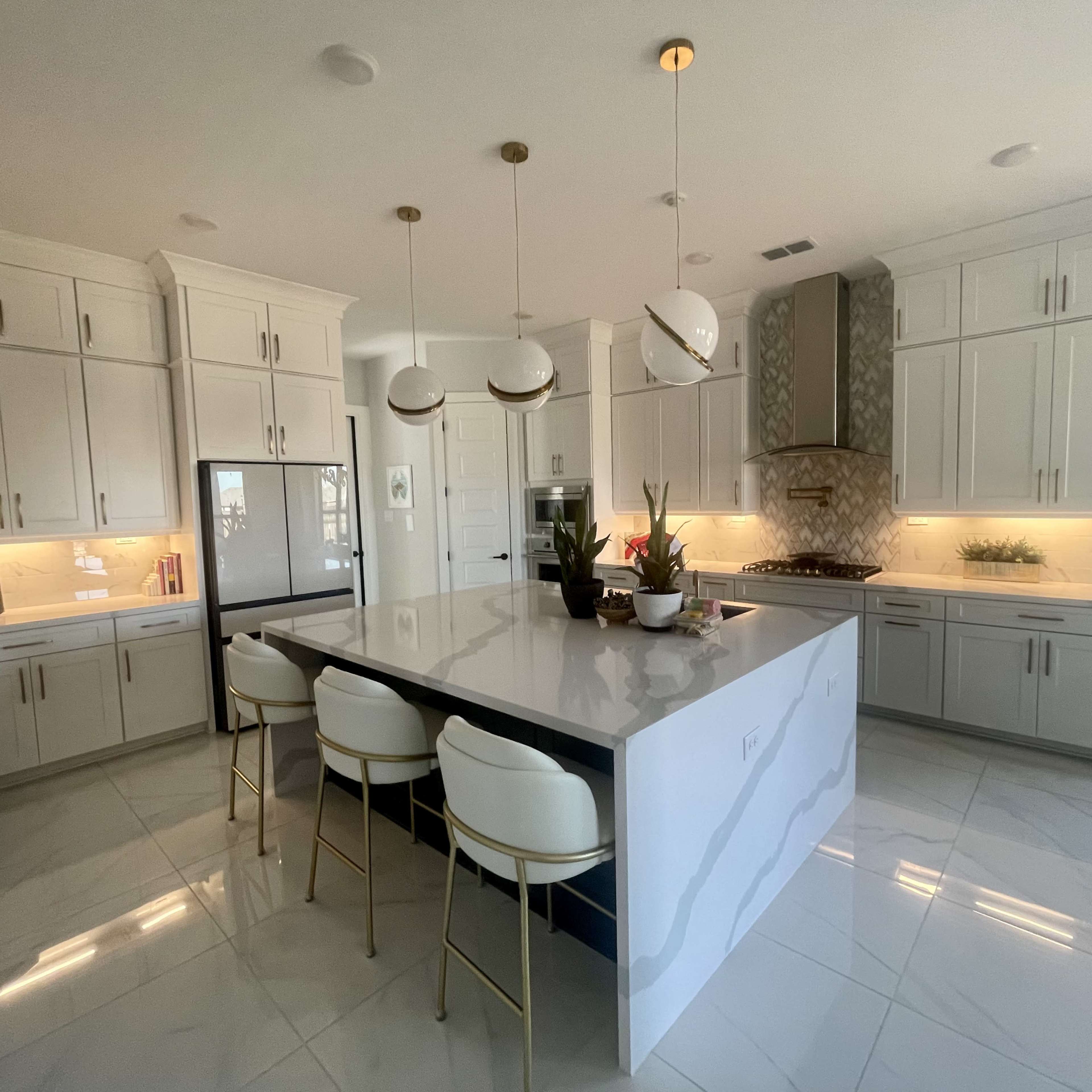 A modern kitchen features a large central island with four stools, white cabinetry, marble countertops, and pendant lighting.