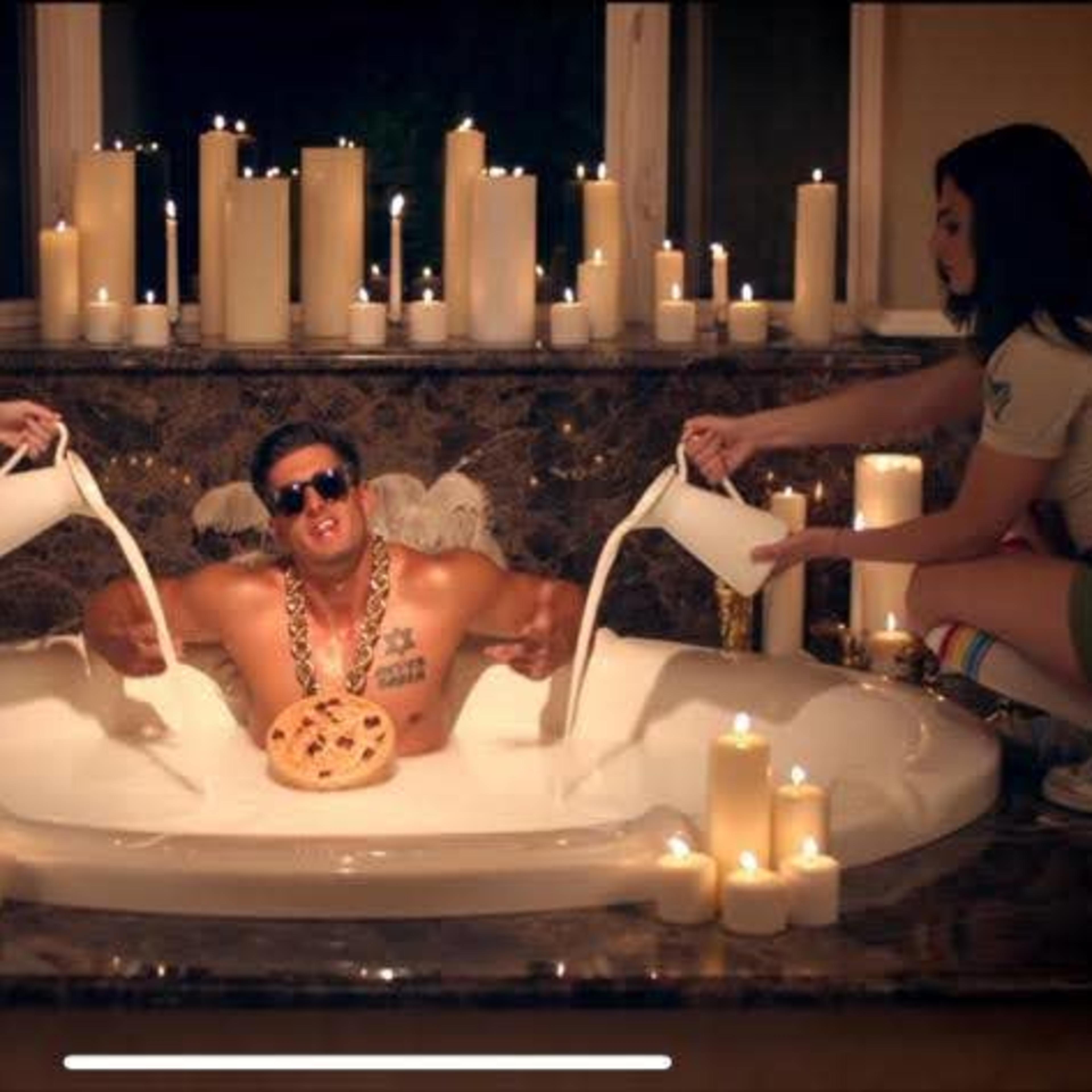 A man wearing sunglasses and a gold chain sits in a large bathtub surrounded by two women, each pouring liquid from buckets while candles illuminate the scene.