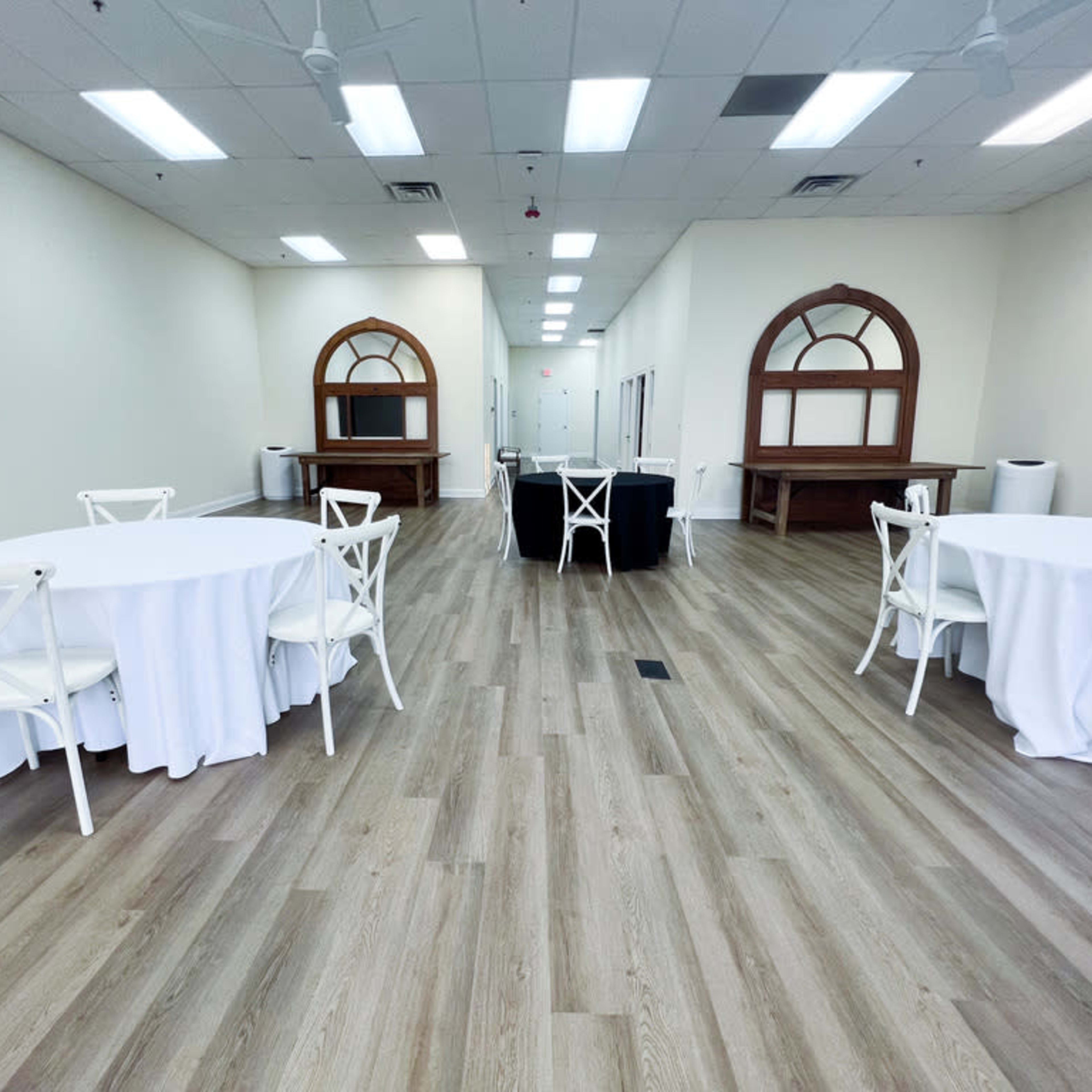 An empty indoor event space with light wooden floors, several white tablecloth-covered round tables, and wooden decor elements at the back.