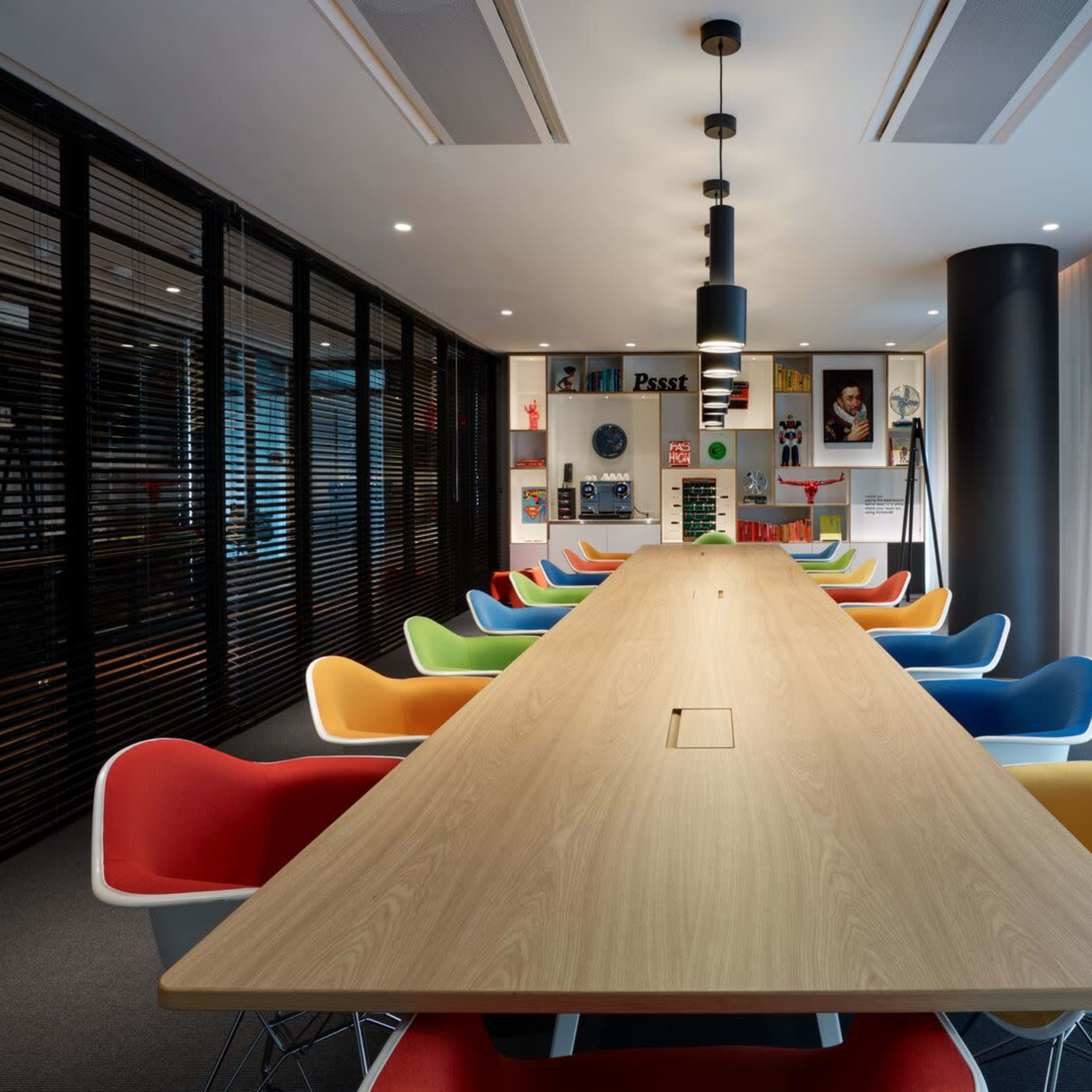 A long wooden table is surrounded by colorful chairs in a modern conference room with large windows and shelves displaying books and decor.
