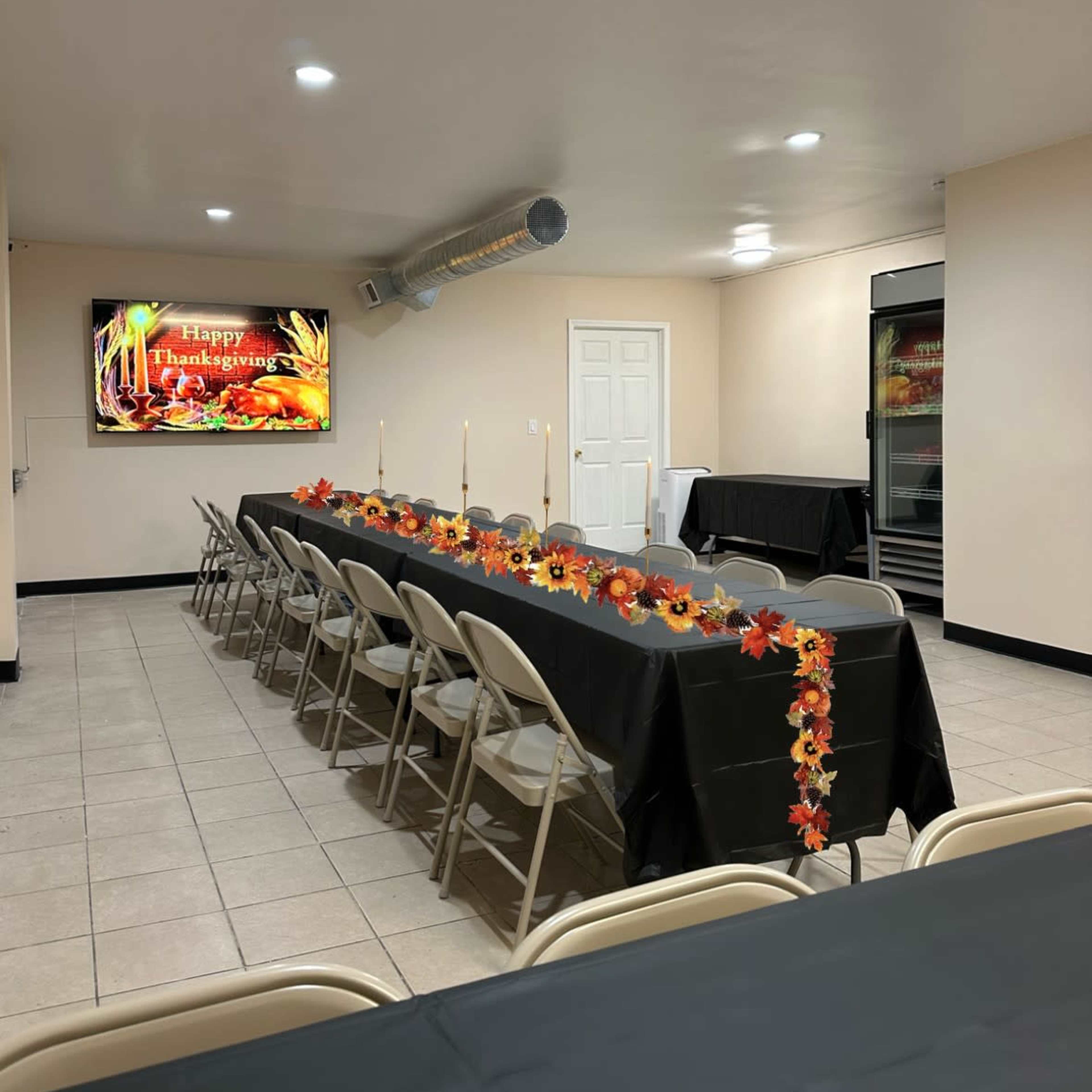 A long table is set in a room with a "Happy Thanksgiving" display on the wall, decorated with a black tablecloth, candles, and autumn-themed decorations.