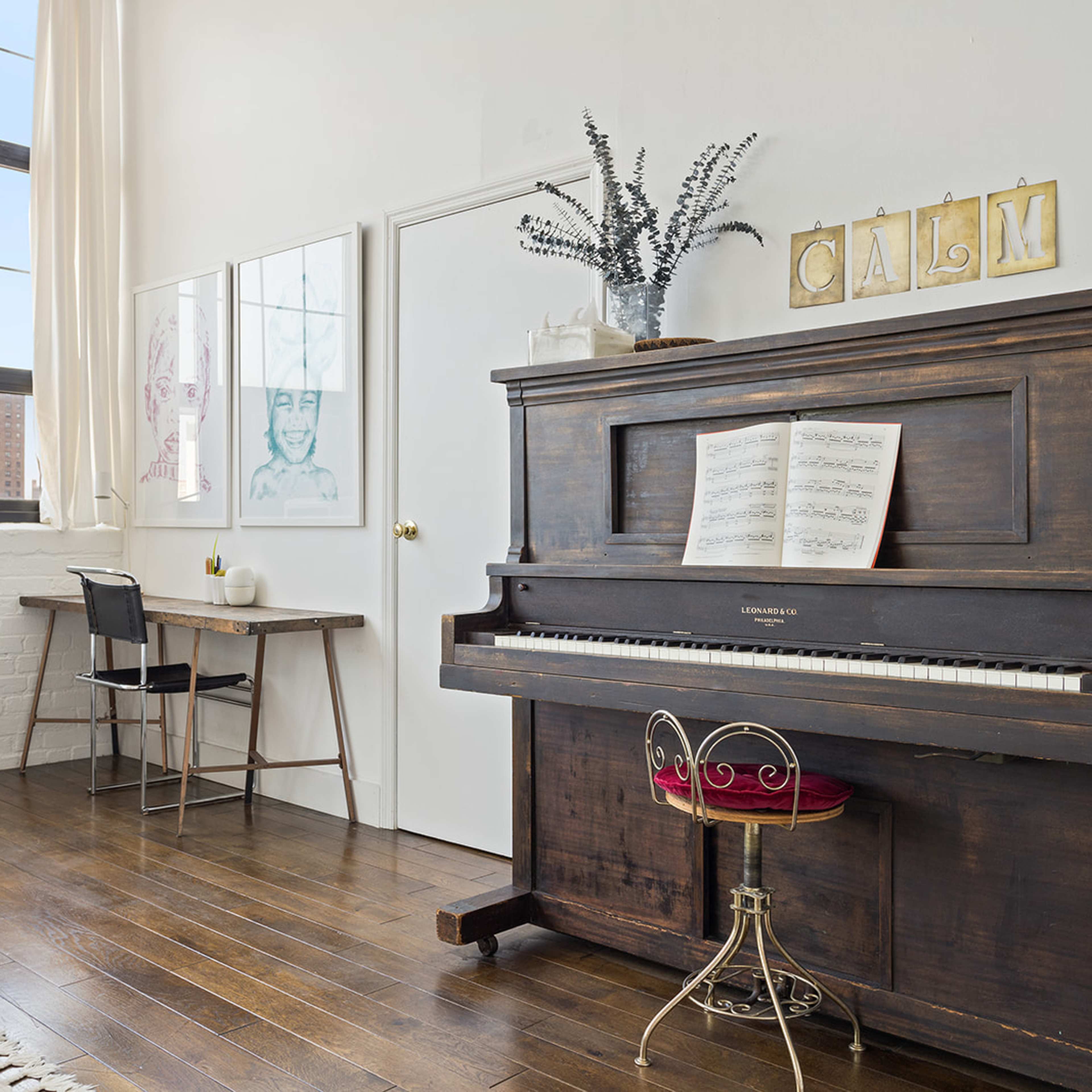 A vintage piano sits beside a small desk in a bright room with large windows and wooden flooring.