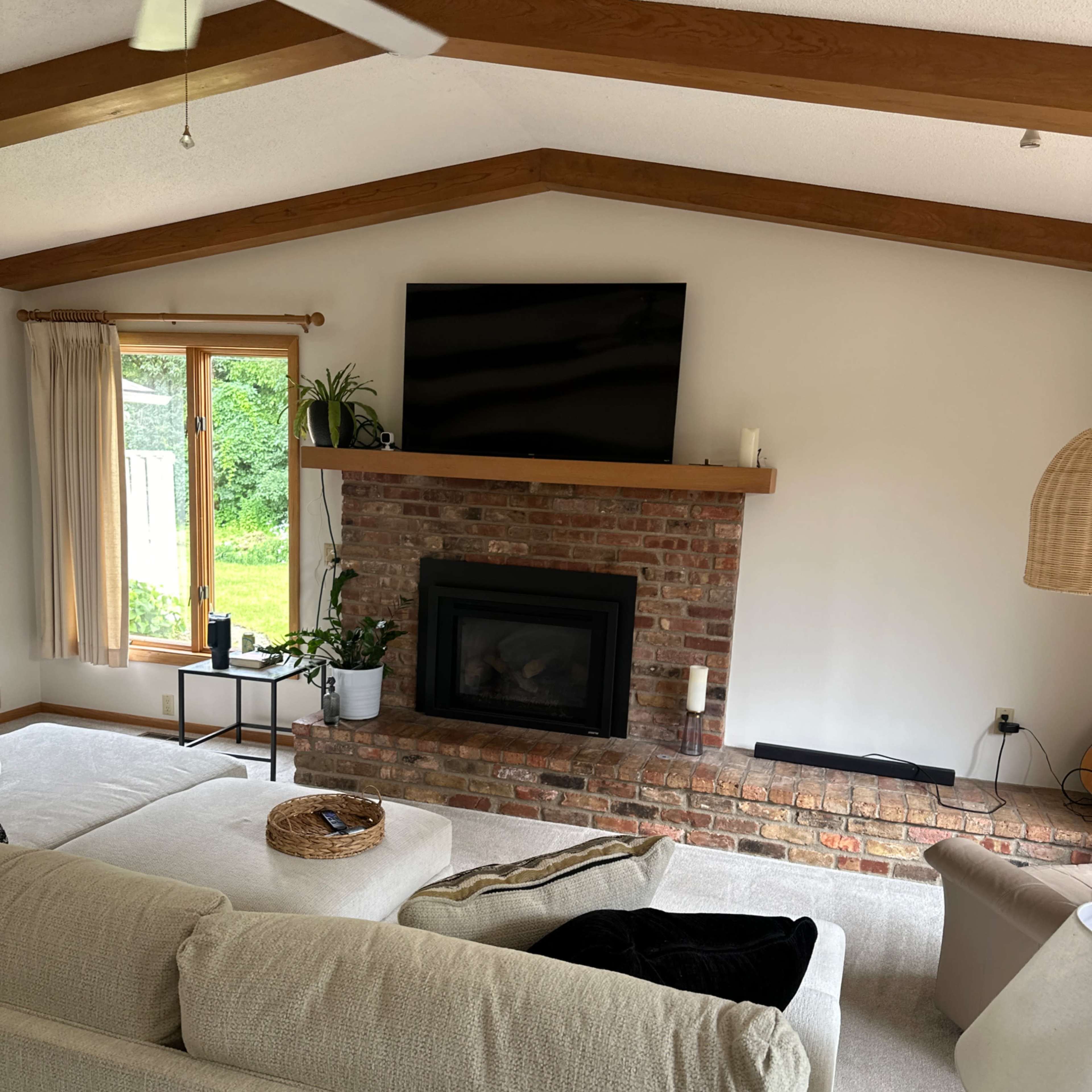 The living room features a brick fireplace with a mounted television above it, a large sectional sofa, and large windows that overlook a green outdoor view.