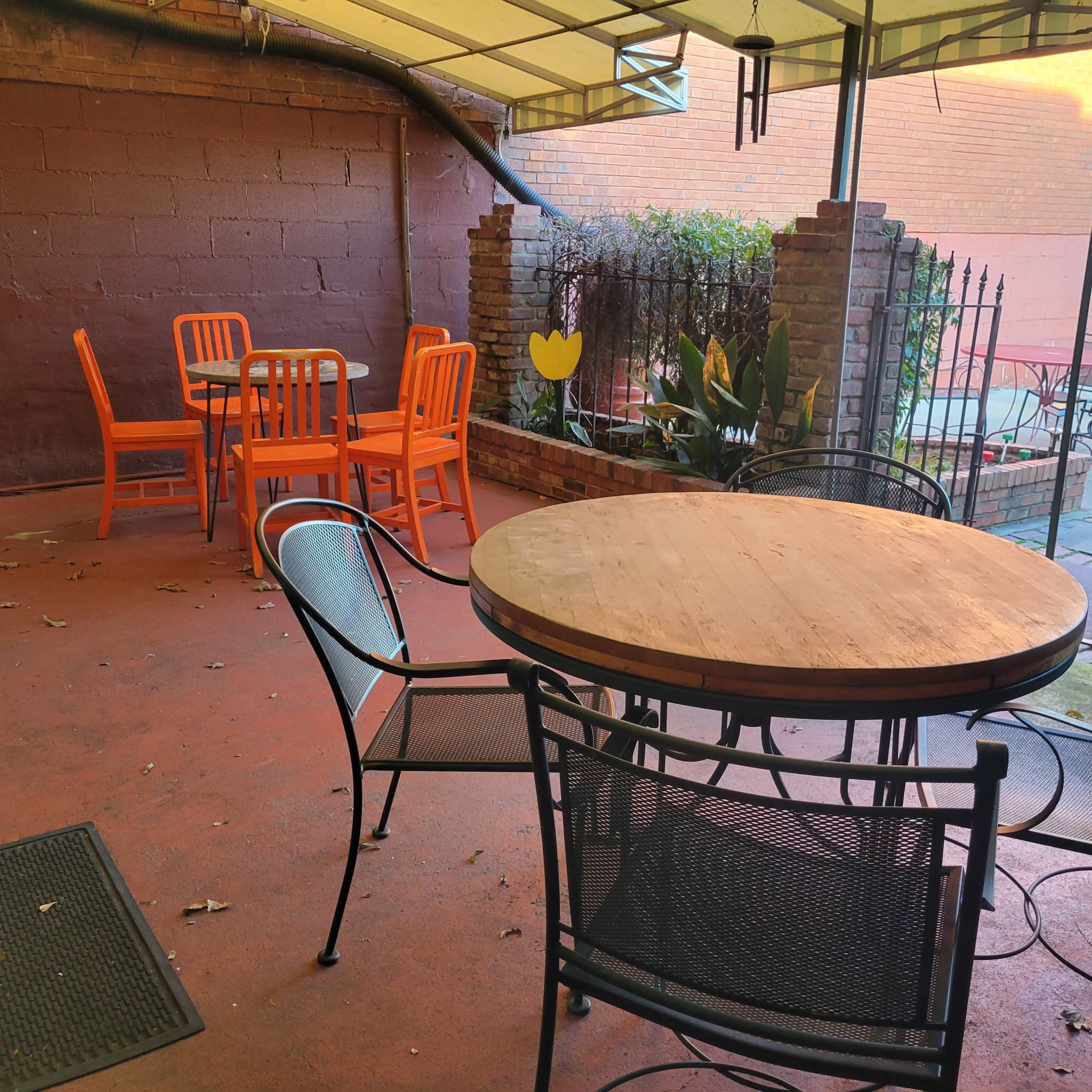 An outdoor seating area features a round wooden table surrounded by black chairs, with orange chairs positioned in the background under a covered patio.