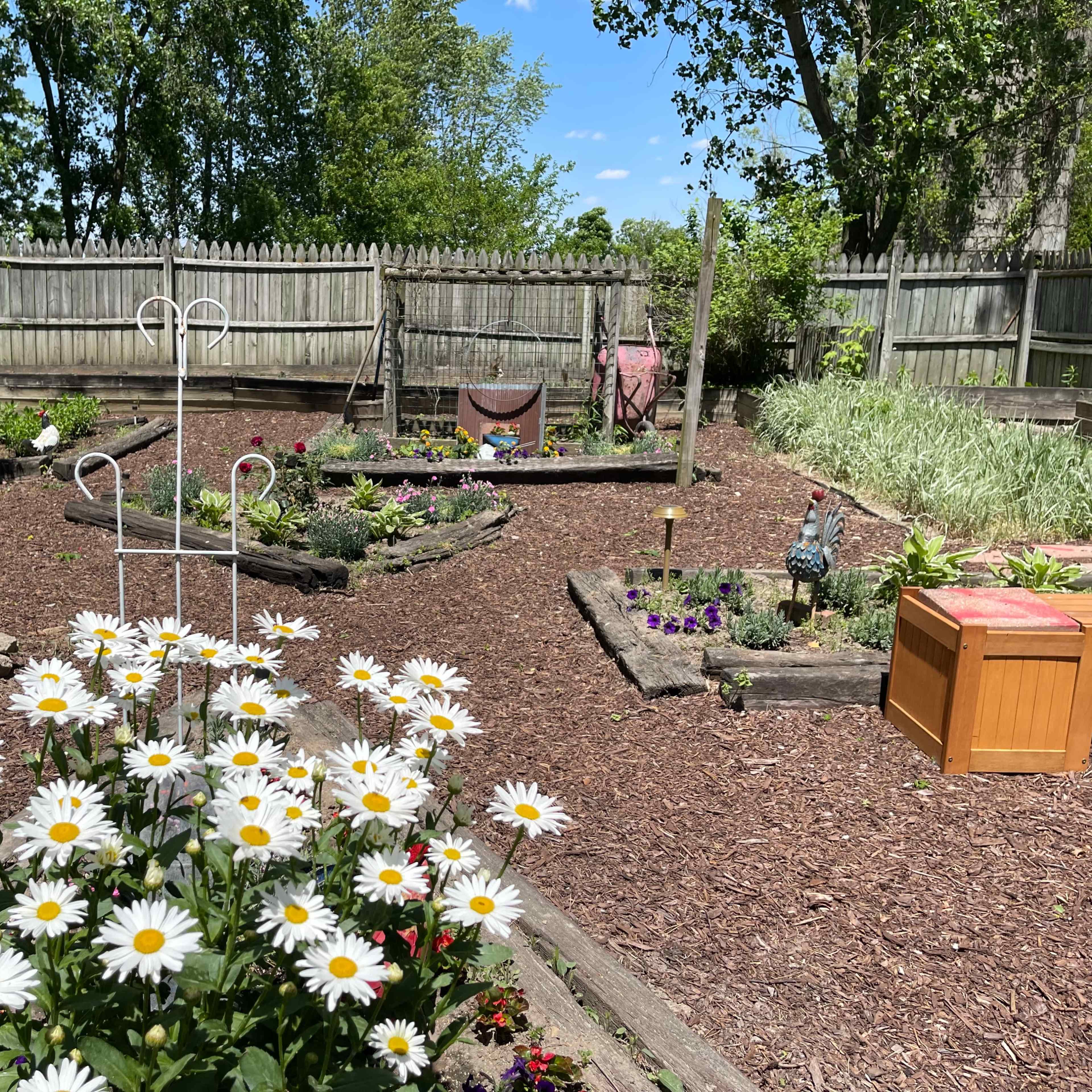 A garden layout with pathways, wooden borders, colorful flowers, and a small wooden storage box under a blue sky.