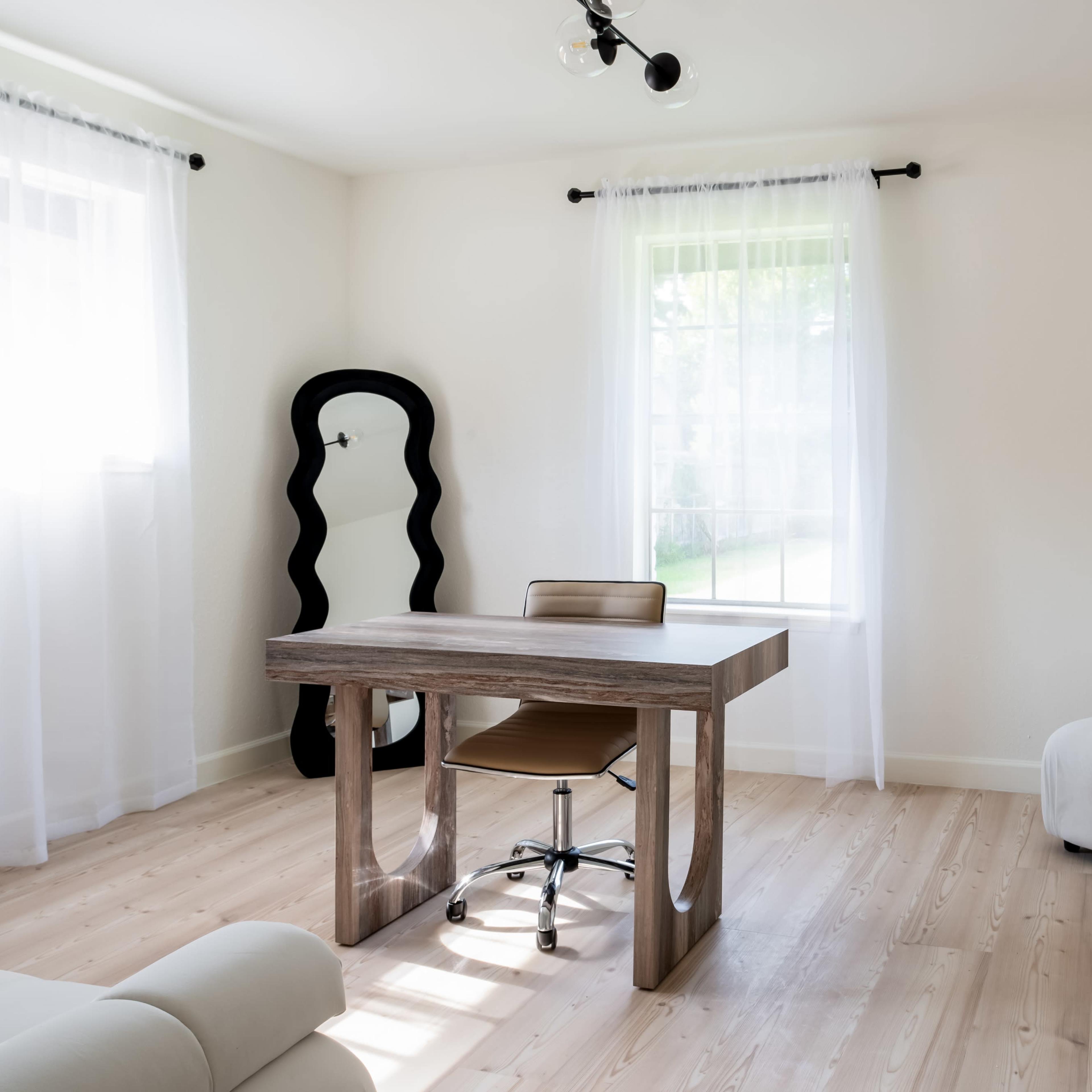 A minimalist room featuring a wooden desk with a chair, a soft couch, and large windows with sheer curtains.