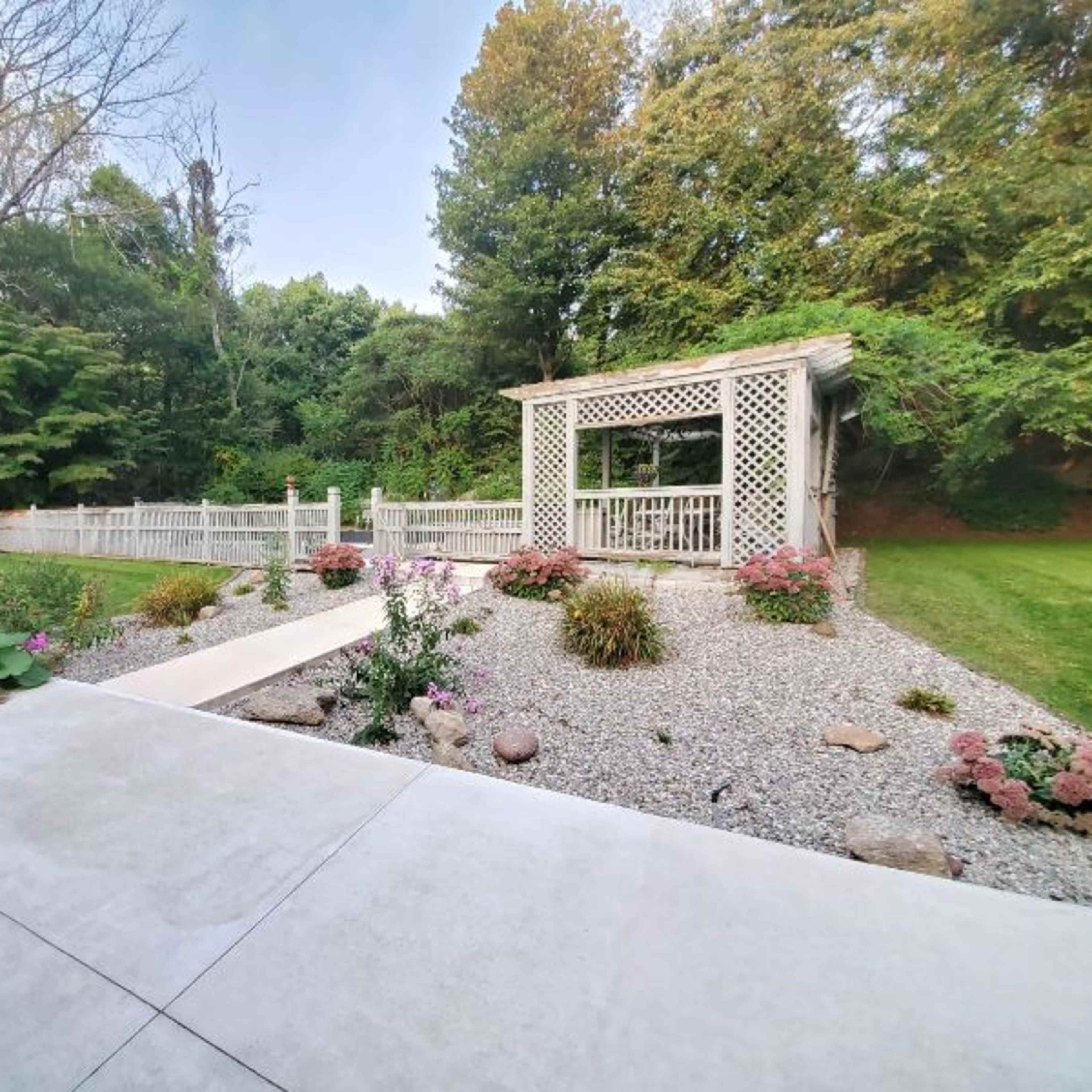 A landscaped garden with a gravel area, colorful flowers, and a gazebo surrounded by greenery.