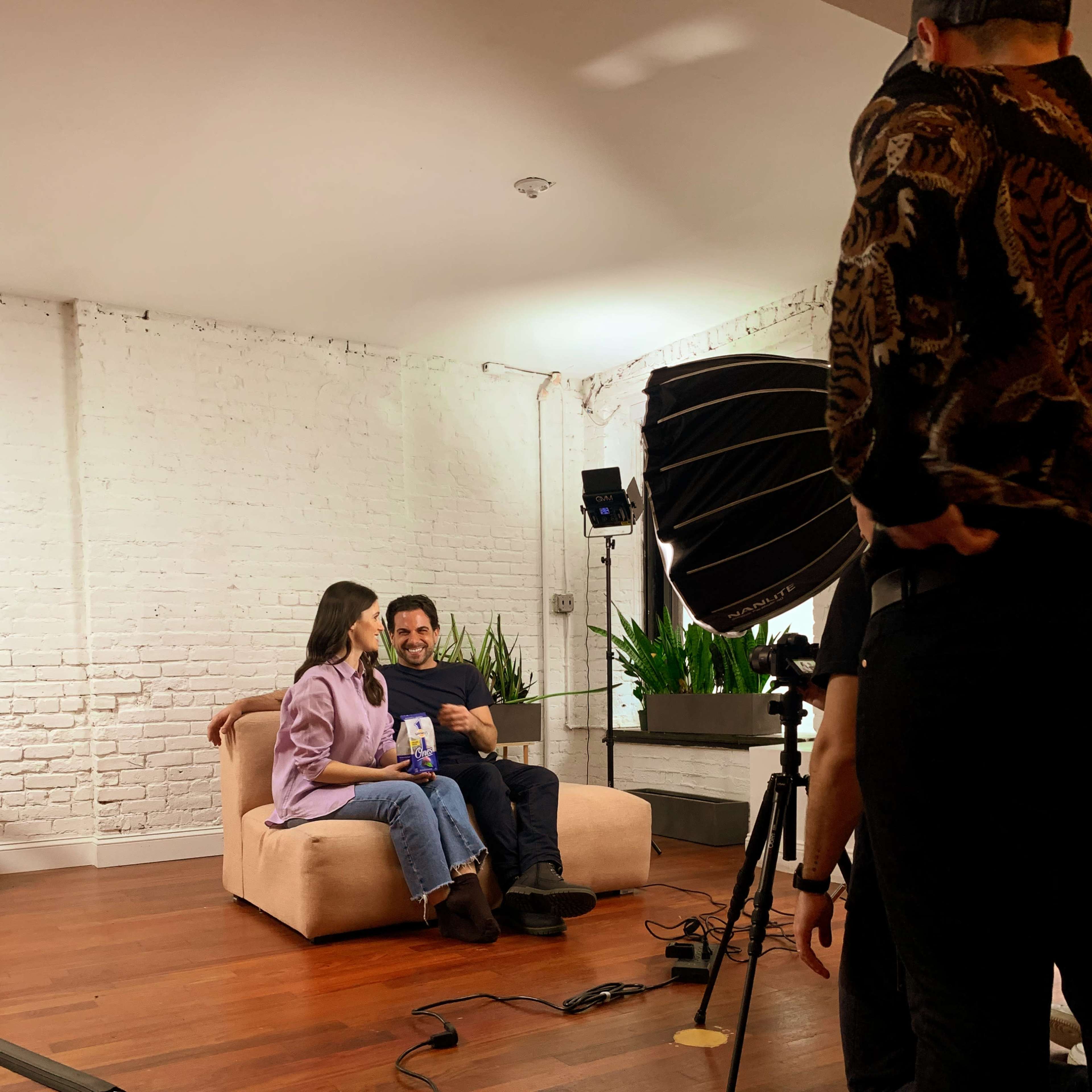 A man and a woman sit on a couch in a bright studio, engaged in conversation while a camera crew prepares to film.
