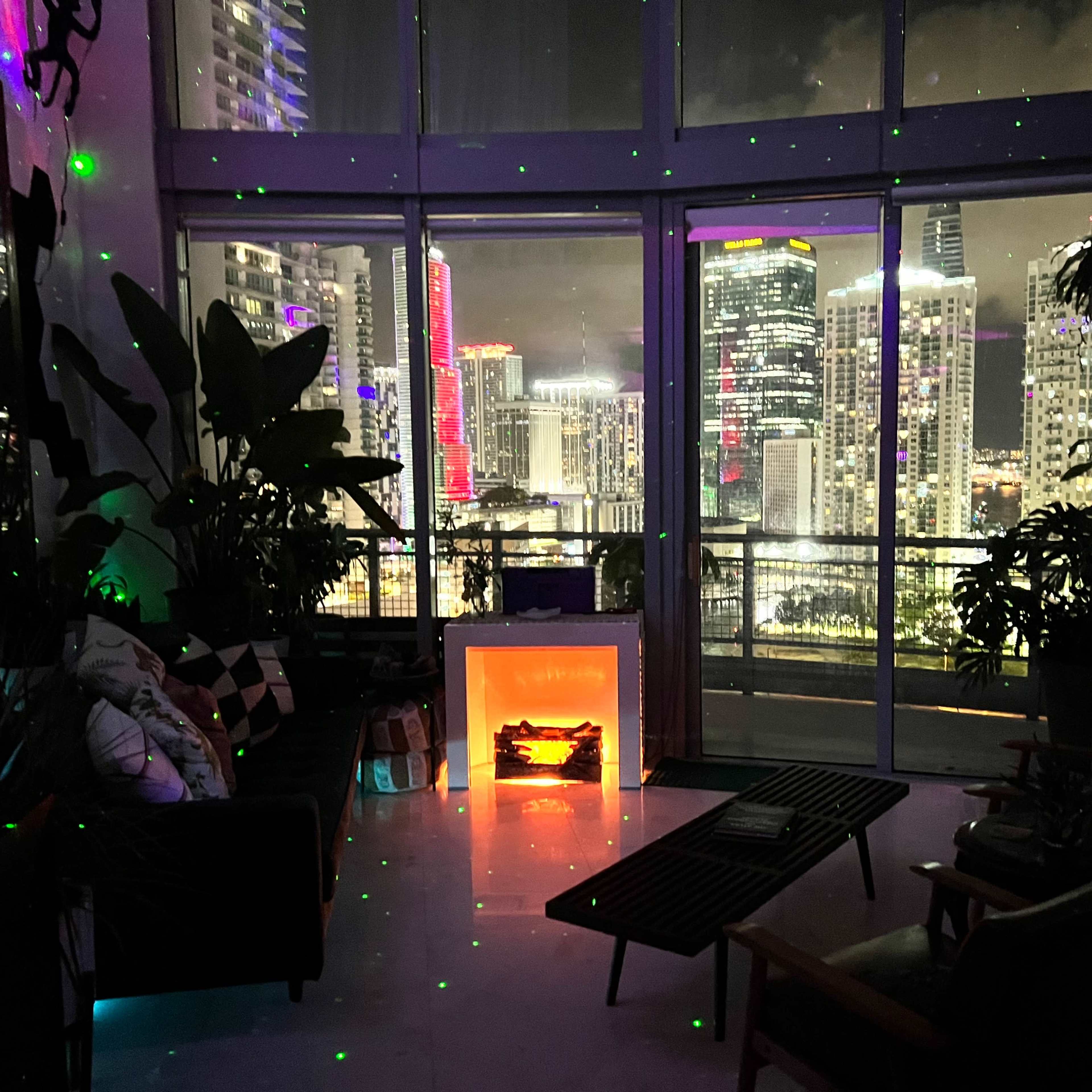 A modern living room features a glowing electric fireplace and large windows overlooking a vibrant city skyline at night.
