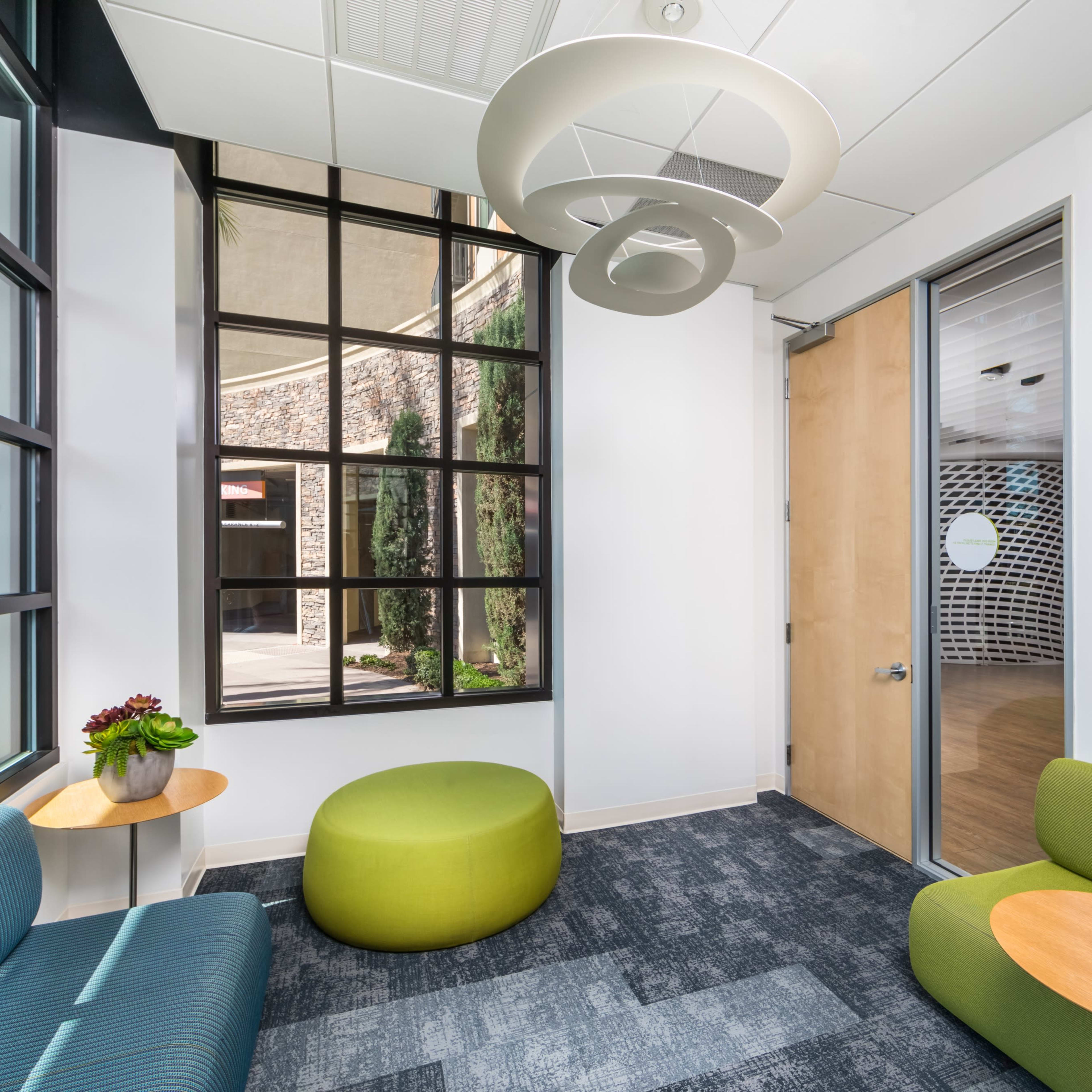 A modern lounge area with two seating options, a round green ottoman, and a small table, surrounded by large windows and a doorway leading to another room.