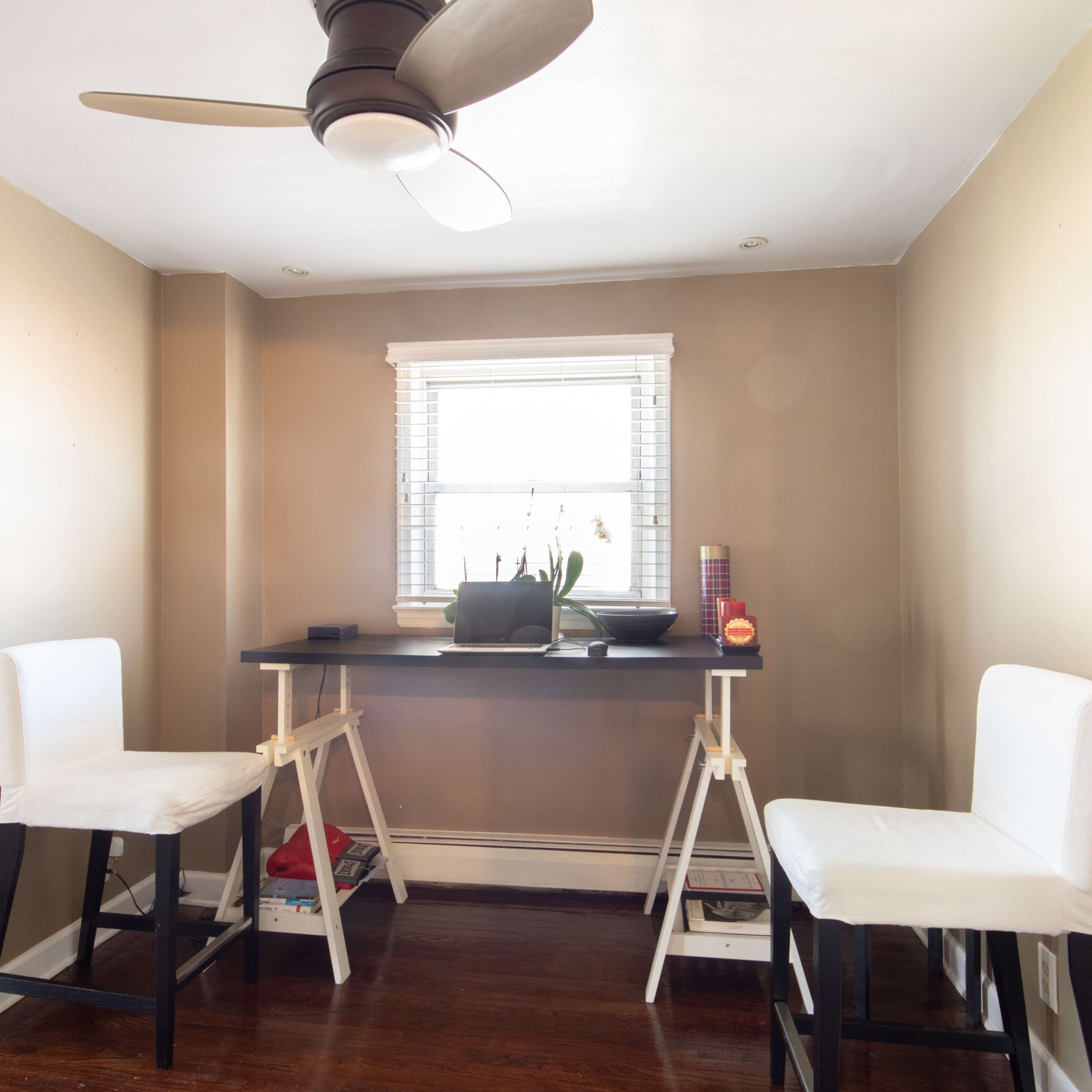 A small room with two white barstools positioned at a narrow table beneath a window with a plant on it.